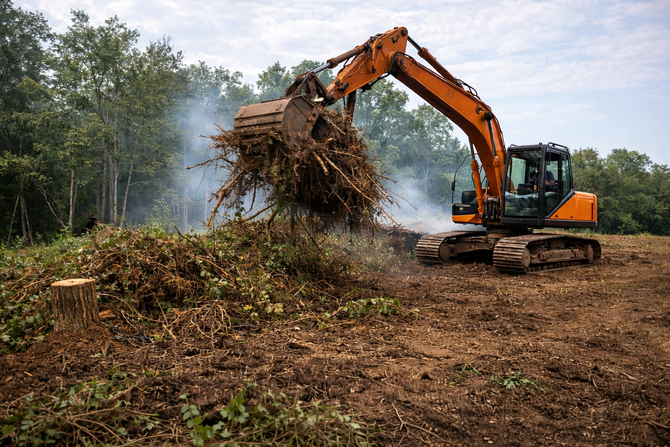 Excavator moving brush in a cleared area, with smoke rising from a pile near trees.