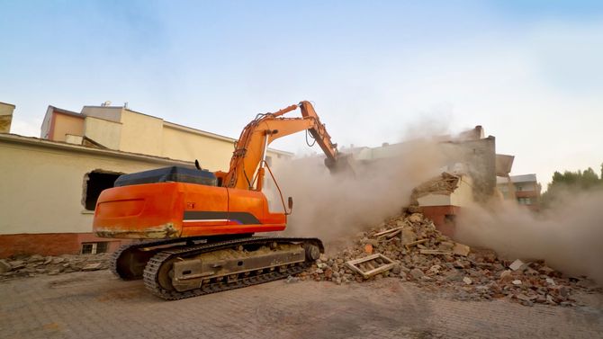 Orange excavator demolishing a building, creating dust and debris.