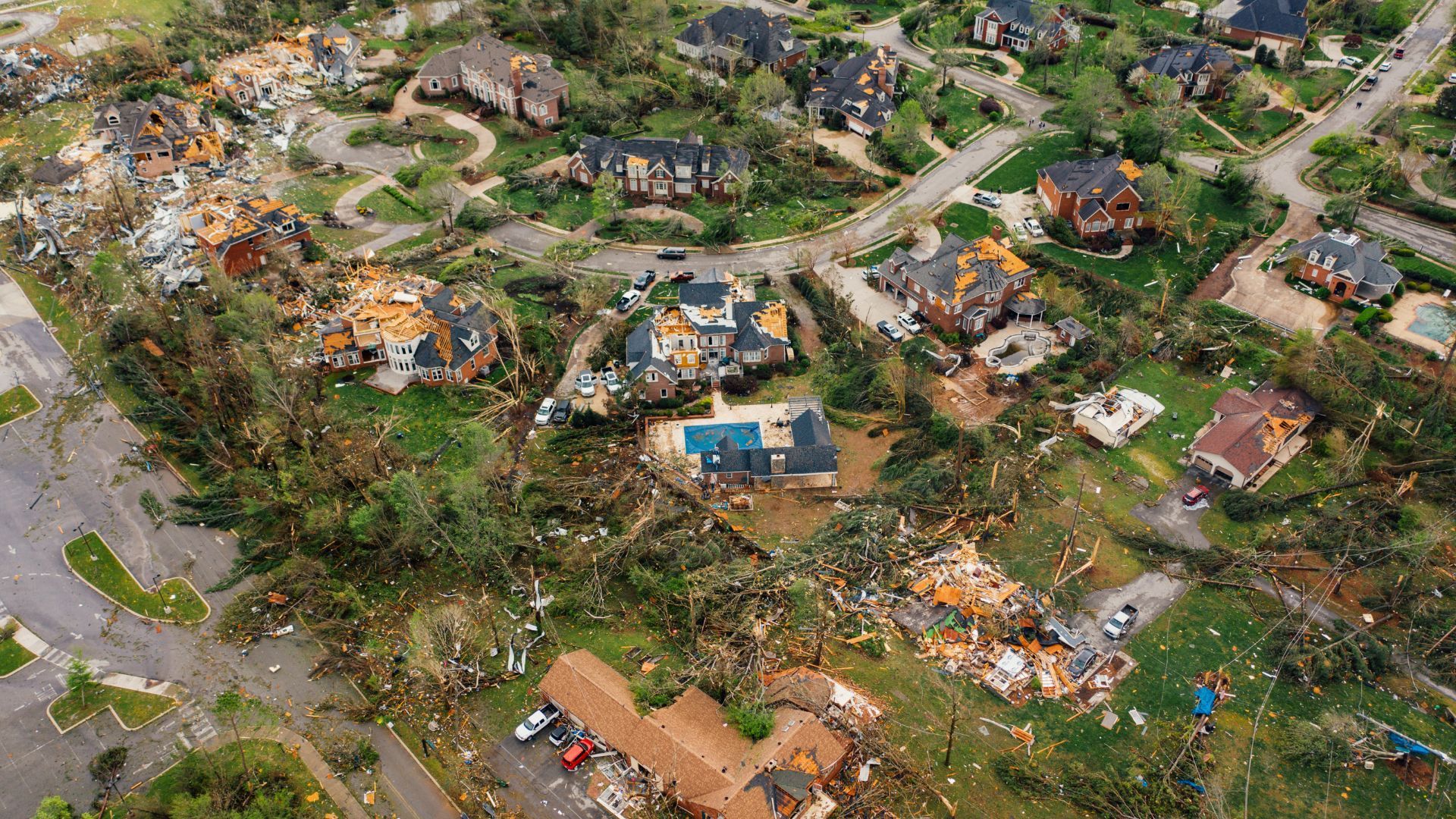 Aerial view of a neighborhood devastated by a tornado. Damaged homes, debris, and fallen trees are scattered across the landscape.
