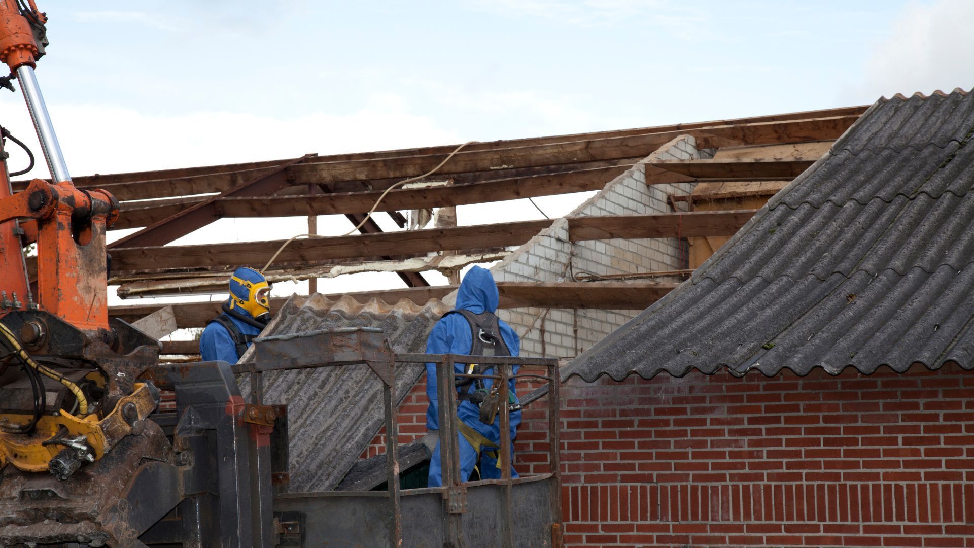 Two workers in blue protective suits operate a lift near a building being demolished, with exposed beams and asbestos roof visible.