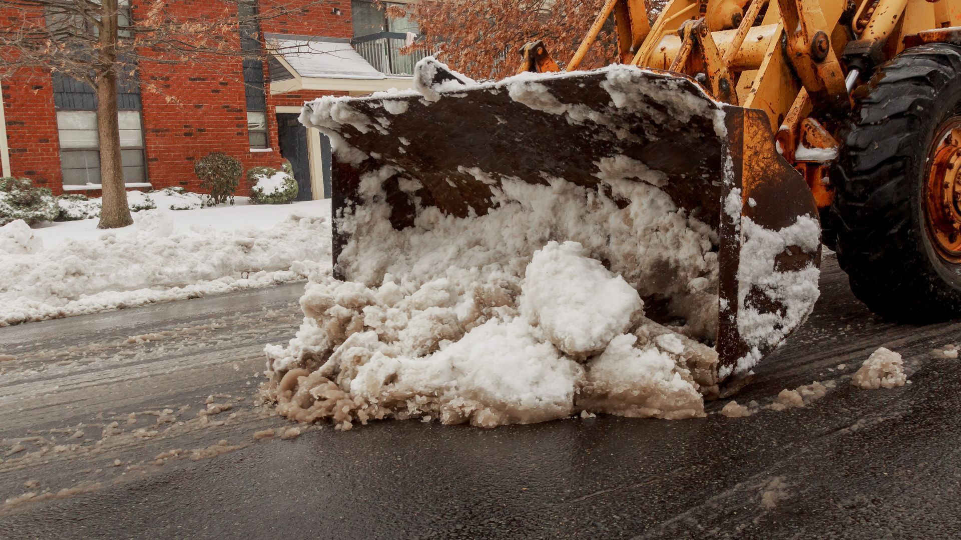Snowplow clearing snow from a street in front of brick buildings.