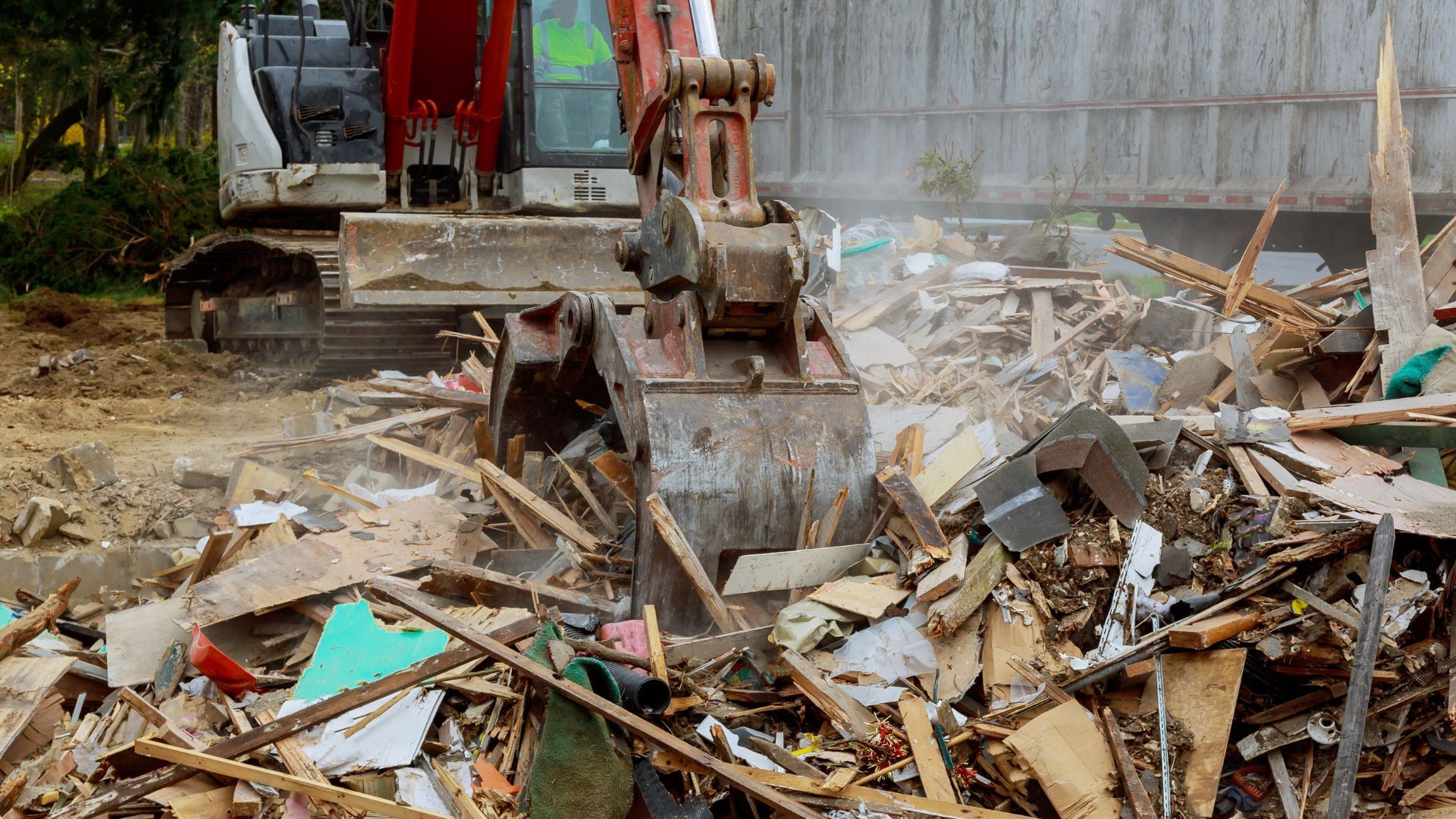 Excavator demolishing a structure, loading debris into a truck; wood, metal, and dust.