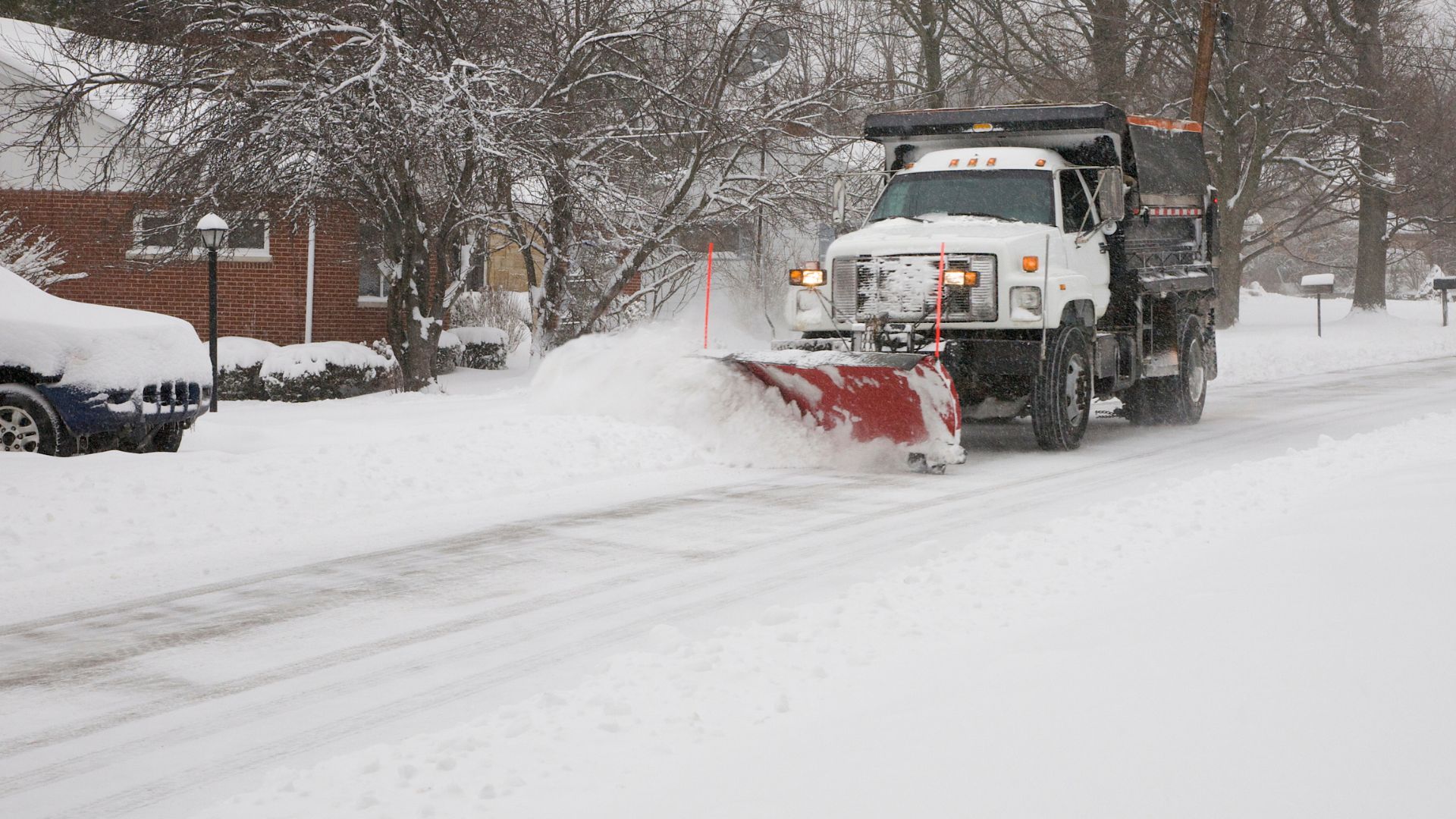 Snowplow clearing snow from a residential street during a snowstorm; a car is parked on the side.