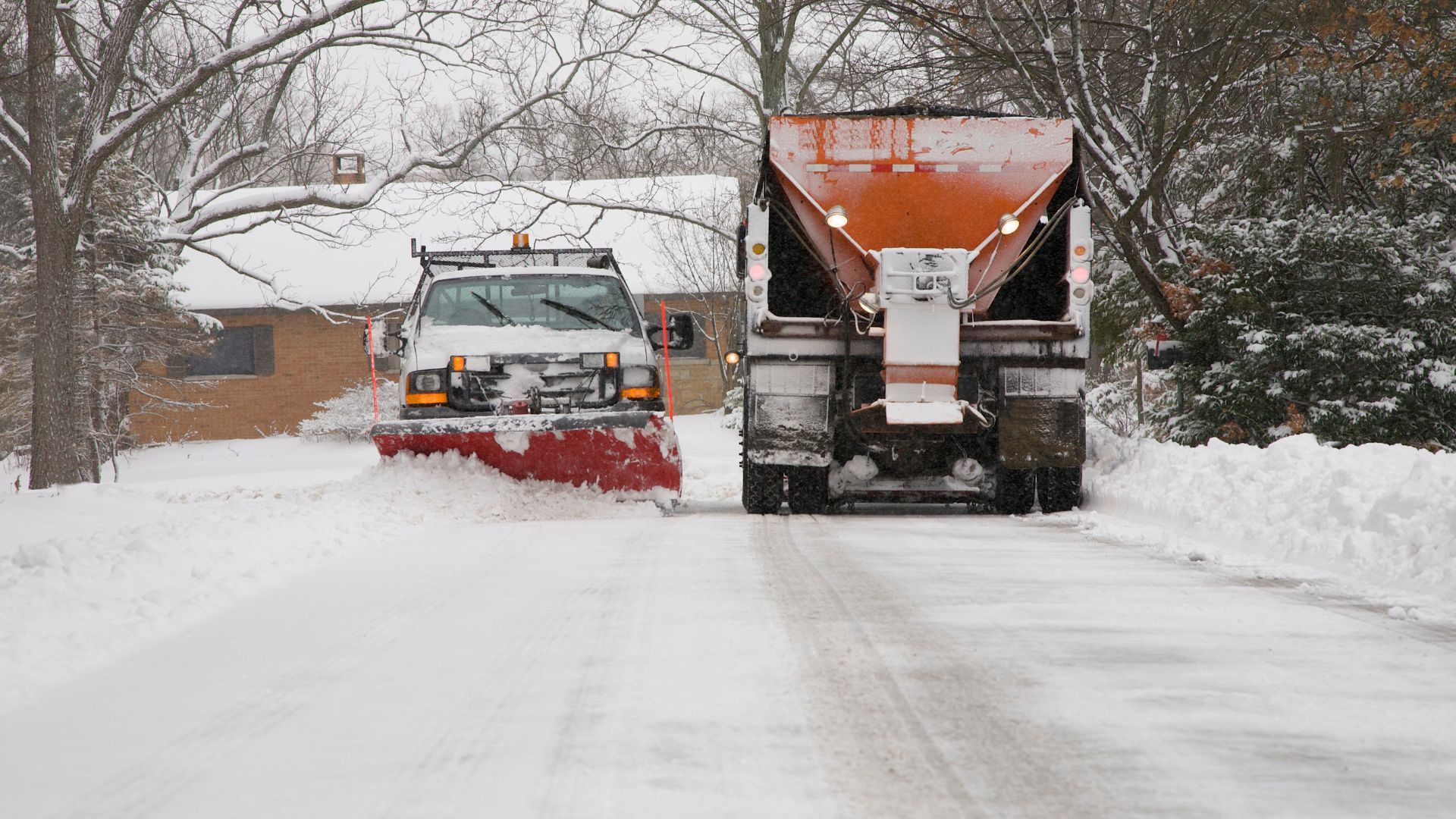 Snowplow and salt truck clearing snow from a residential road during a snowfall.