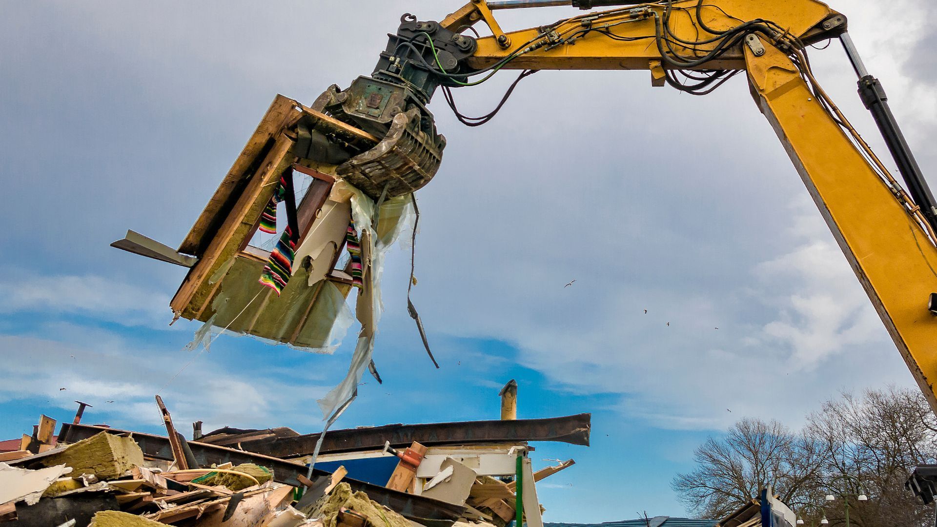 Excavator demolishes a building, holding debris against a cloudy sky.