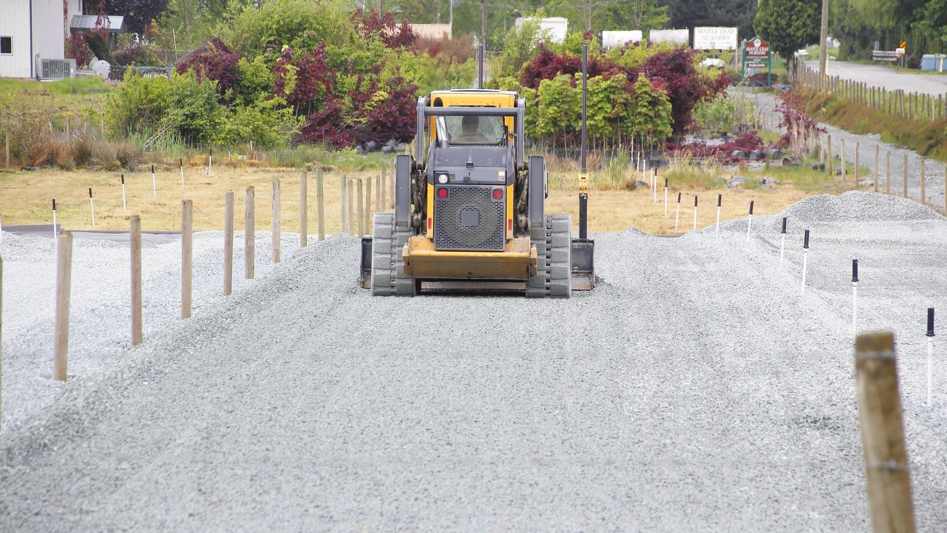 A yellow construction vehicle leveling gravel at a construction site. Wooden stakes line the perimeter.