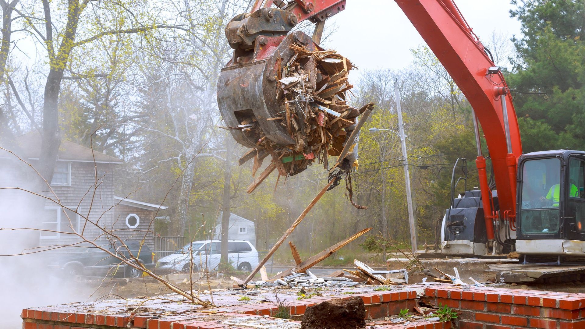 Red excavator demolishing a brick structure, lifting debris in a grapple. Misty atmosphere, rural setting.