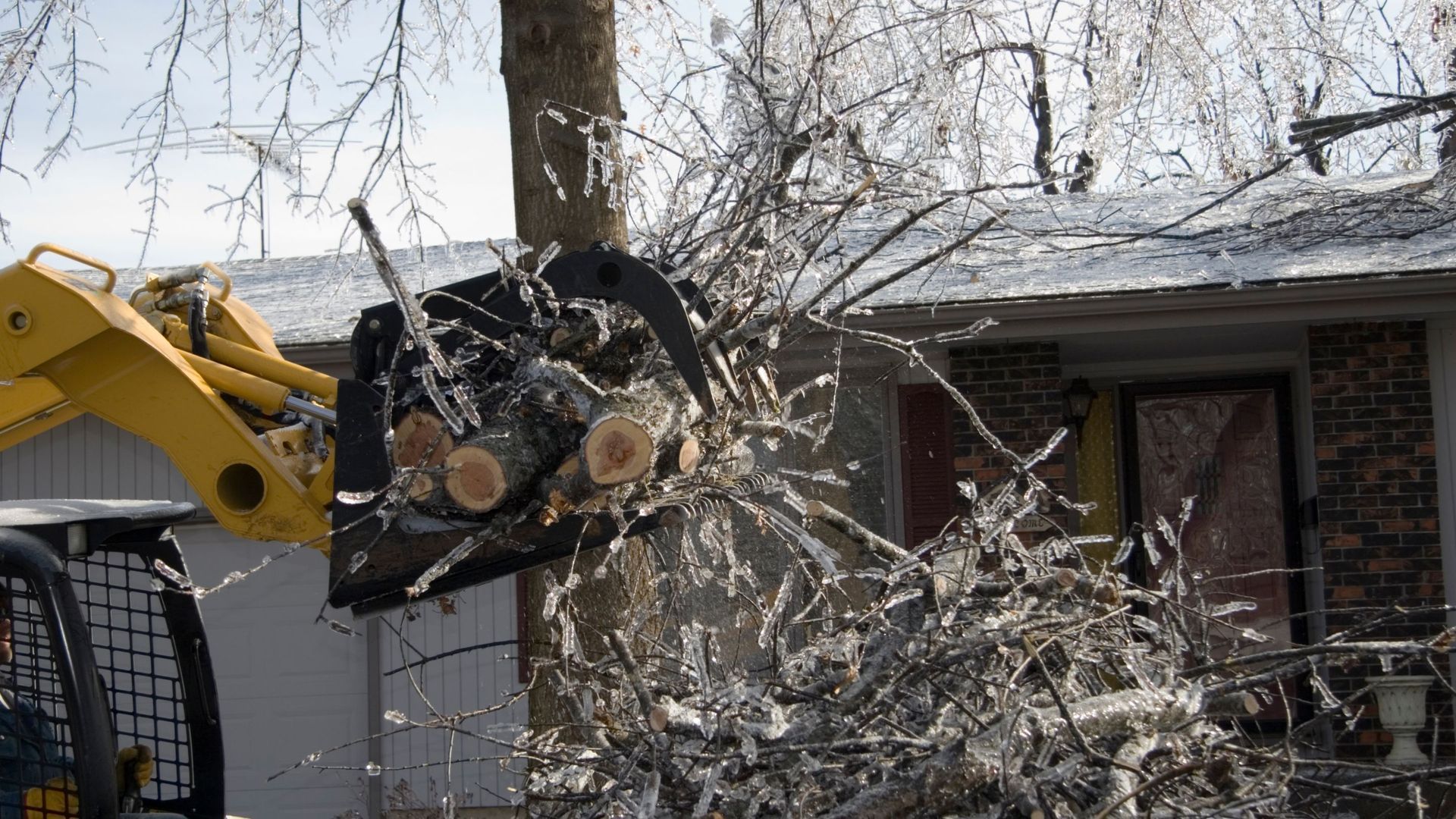 Yellow skid steer carrying tree branches near a house with ice-covered roof.