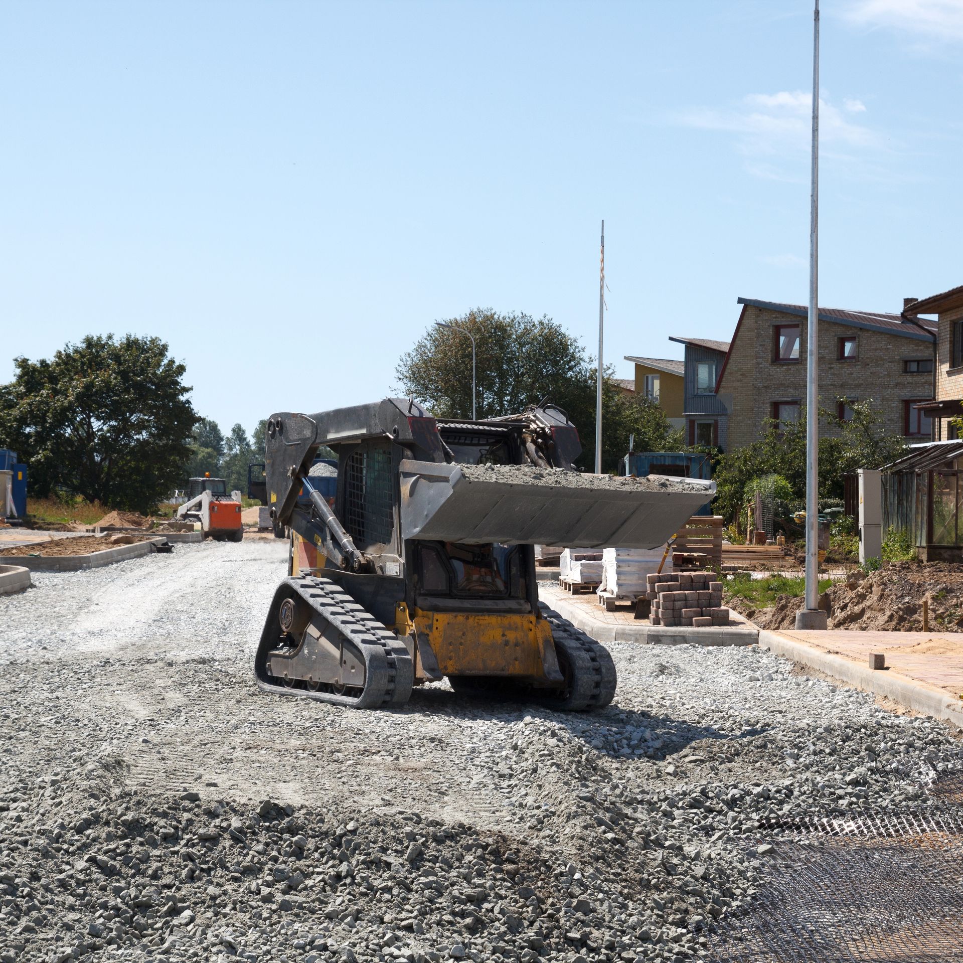 A small, yellow construction vehicle spreading gravel on a road under construction near homes.