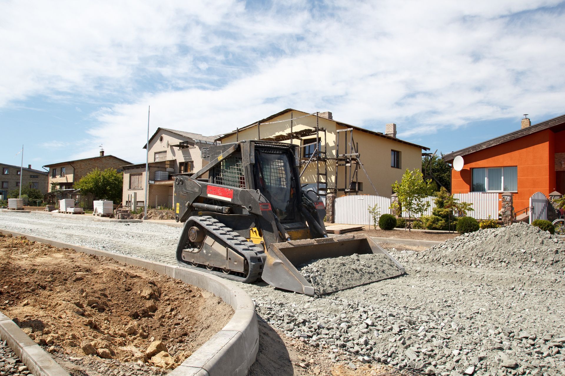 Skid steer loader on a gravel road, working on construction near houses under a blue sky.