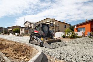 Skid steer loader on a gravel road, working near houses under a blue sky.