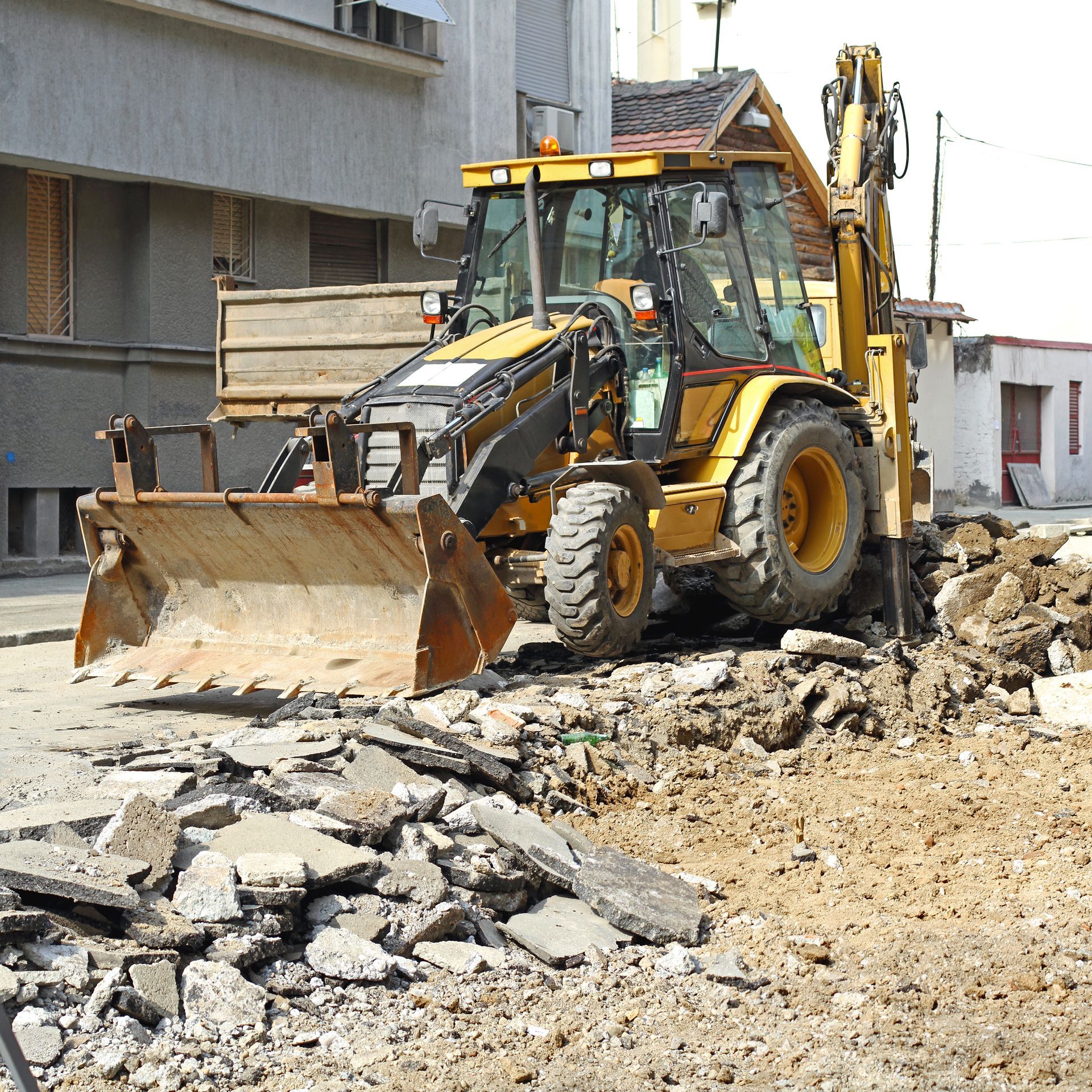 Yellow backhoe digging in a construction site, asphalt debris in foreground, building in background.