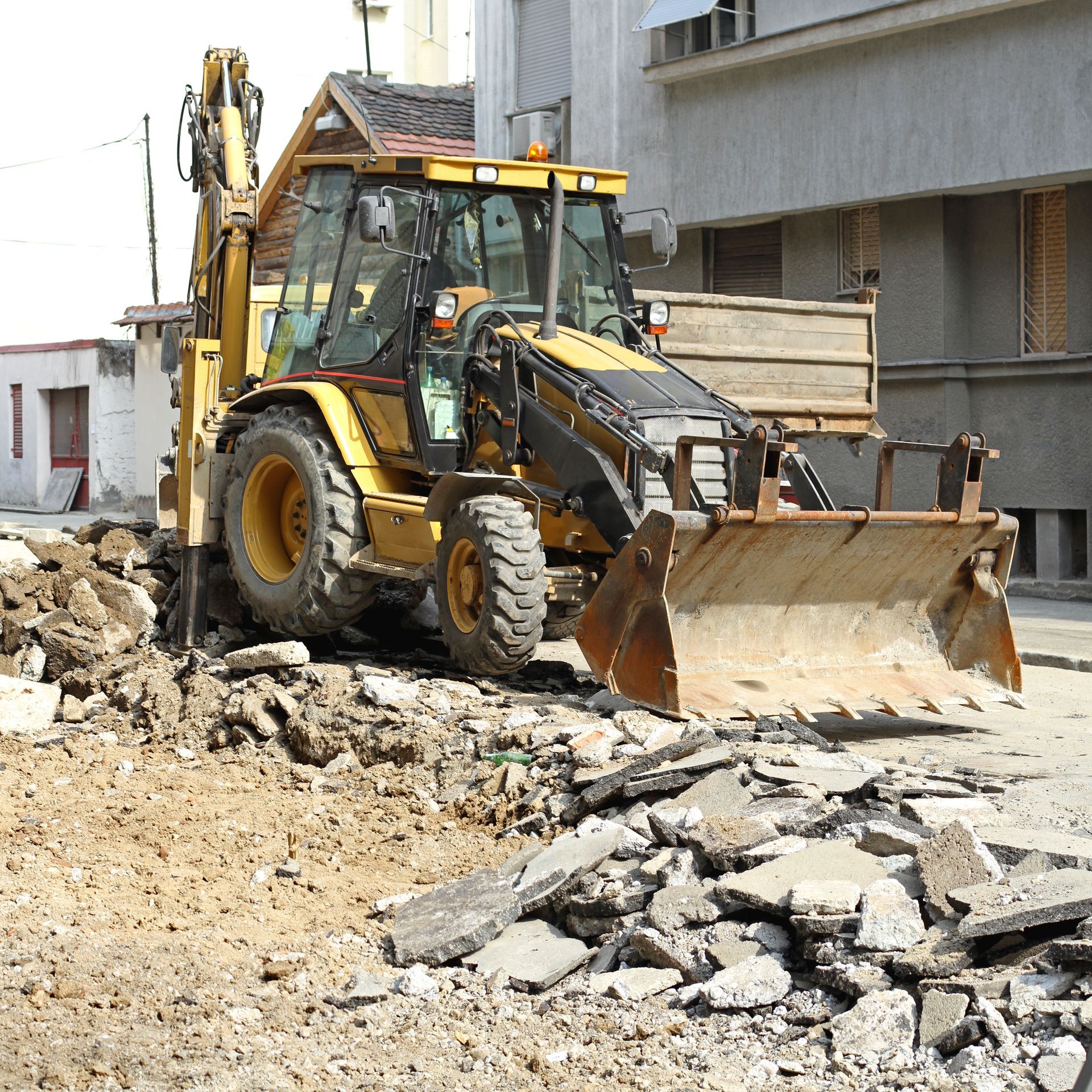 Yellow backhoe on a construction site, scooping up debris near a building.