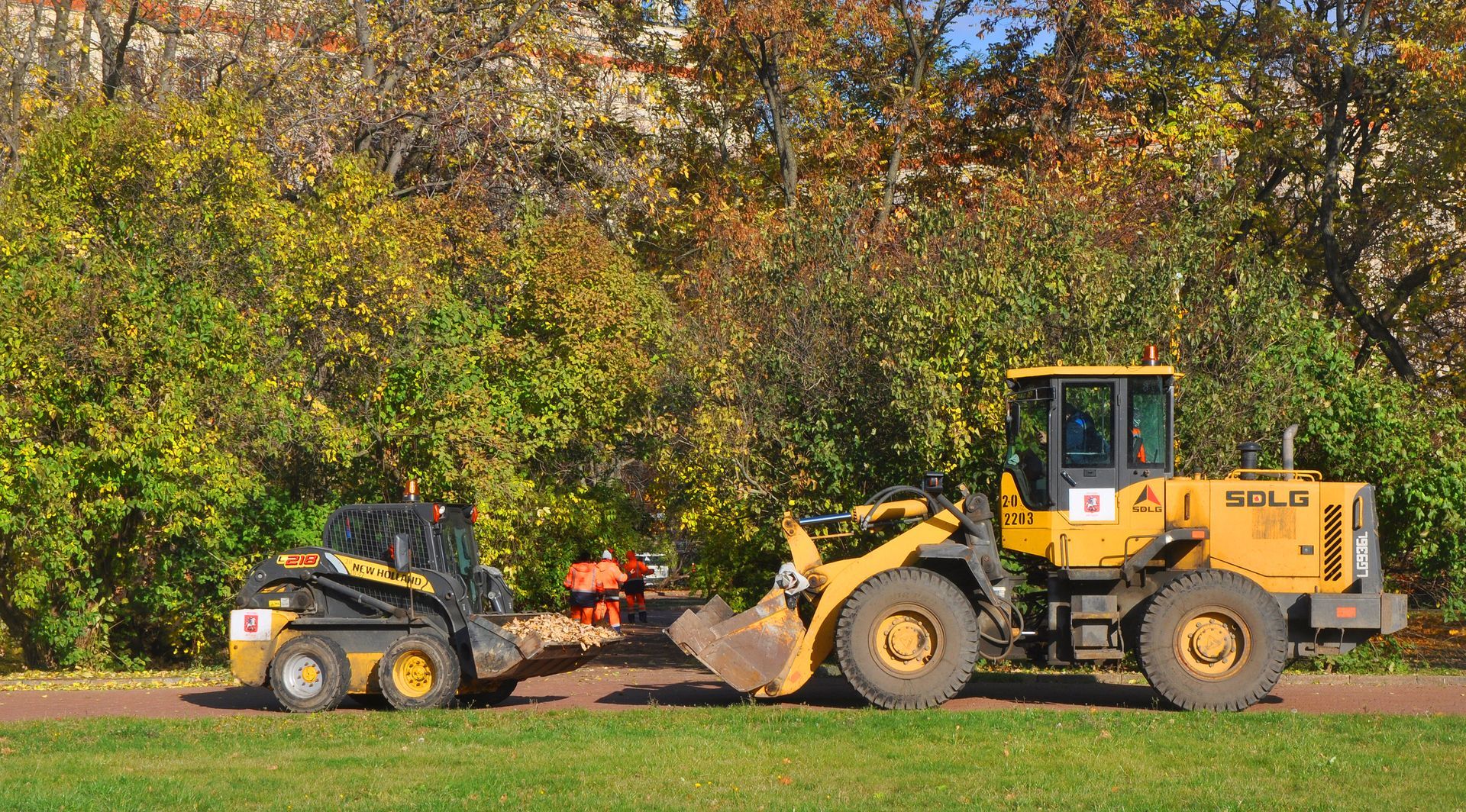 Yellow front-end loader towing a small skid steer on grass, trees in the background.