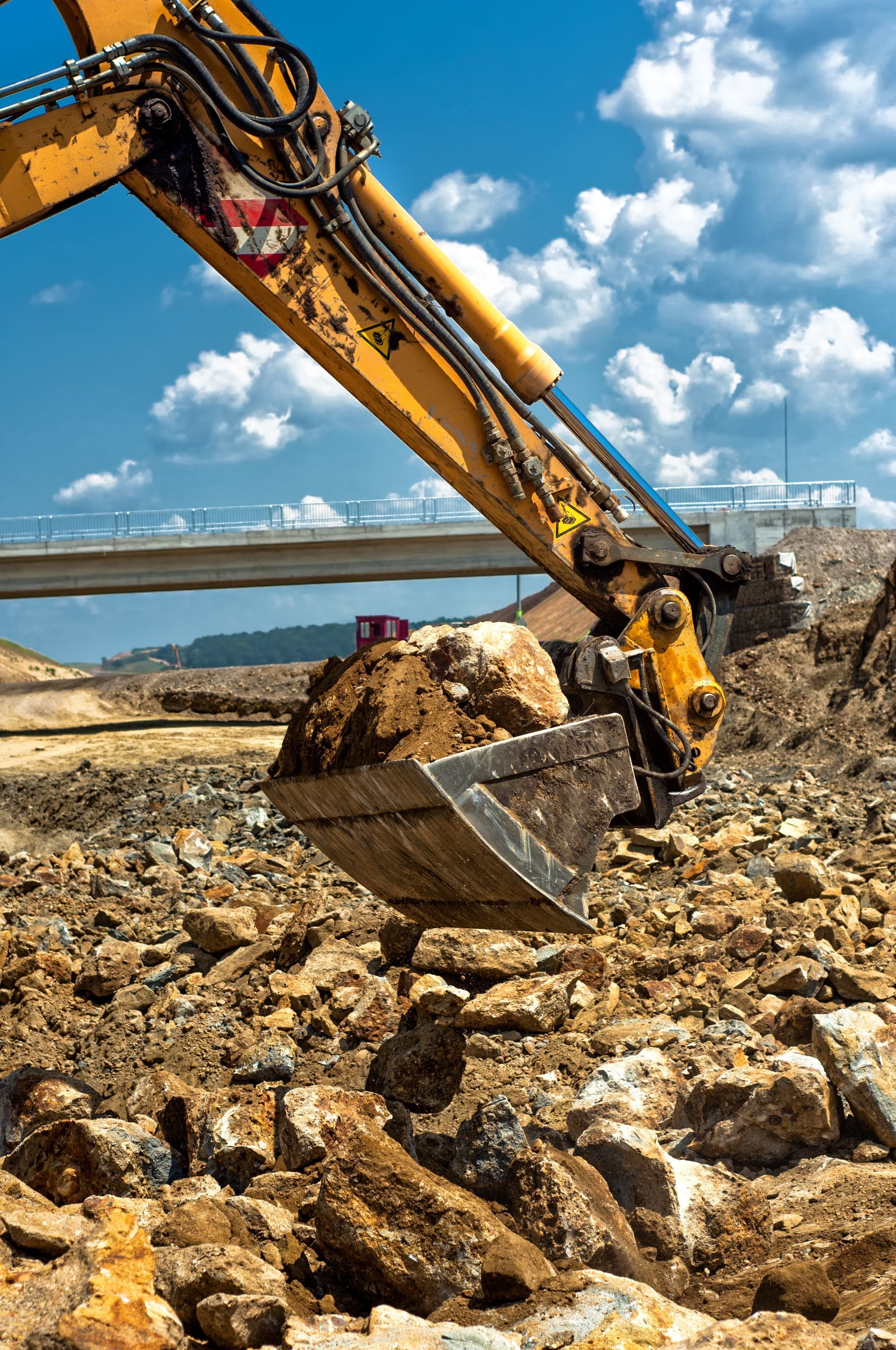 Yellow excavator bucket holding dirt at a construction site with bridge in the background.