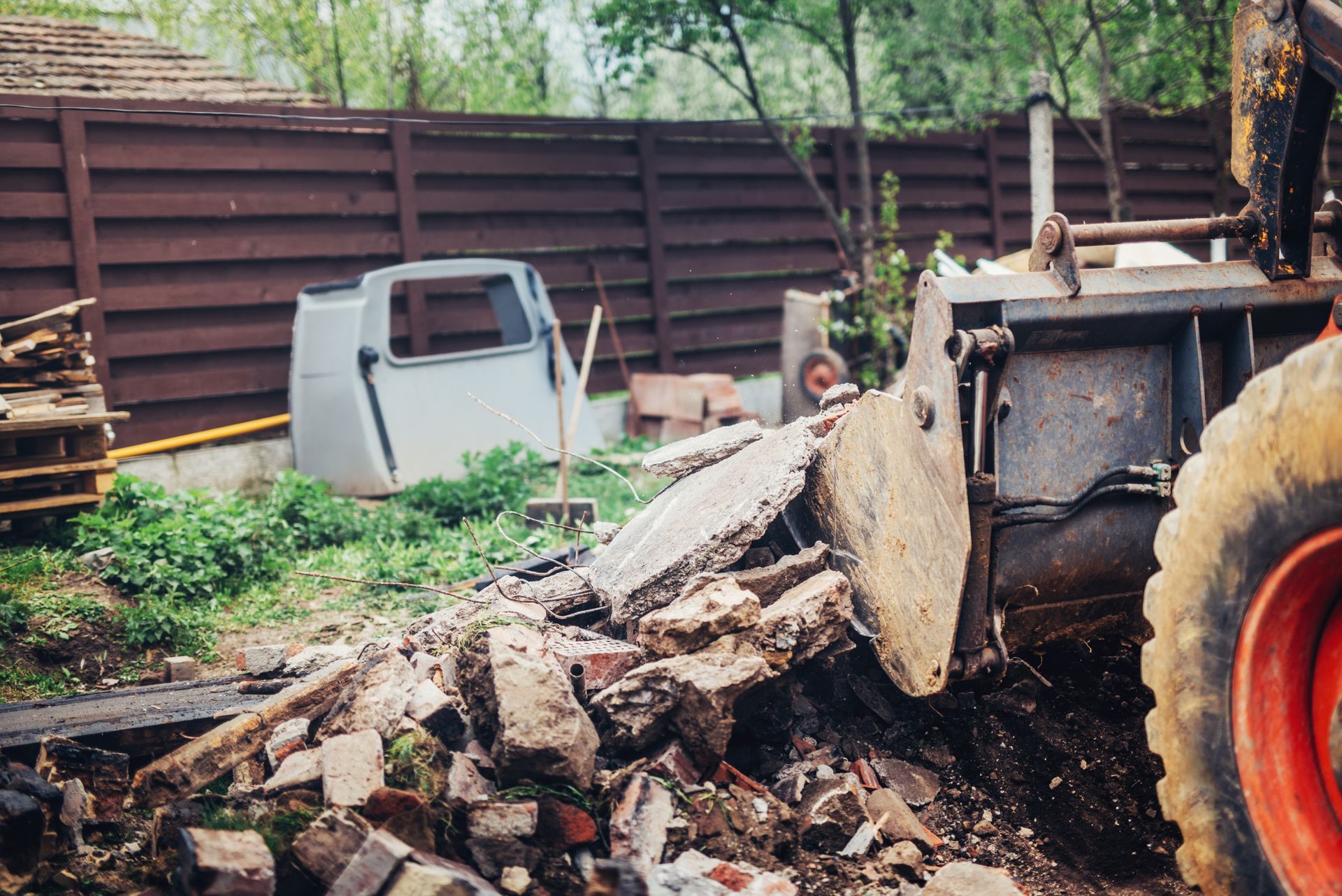 Front-end loader removing debris; bricks, concrete, and cab in a backyard. Brown fence and greenery in the background.
