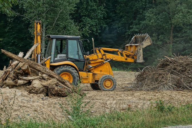 Yellow backhoe clearing debris in a wooded area.