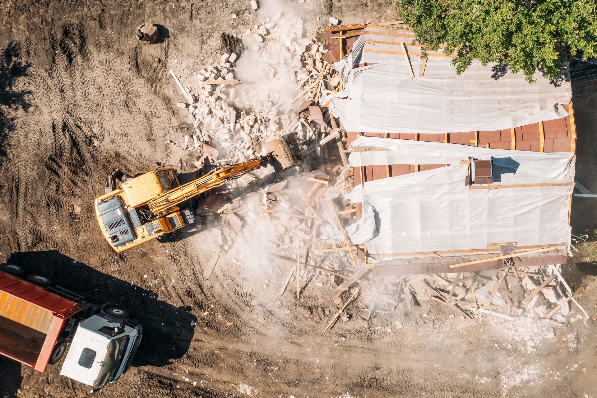 Overhead view of a yellow excavator demolishing a building; a truck is nearby on a dusty worksite.