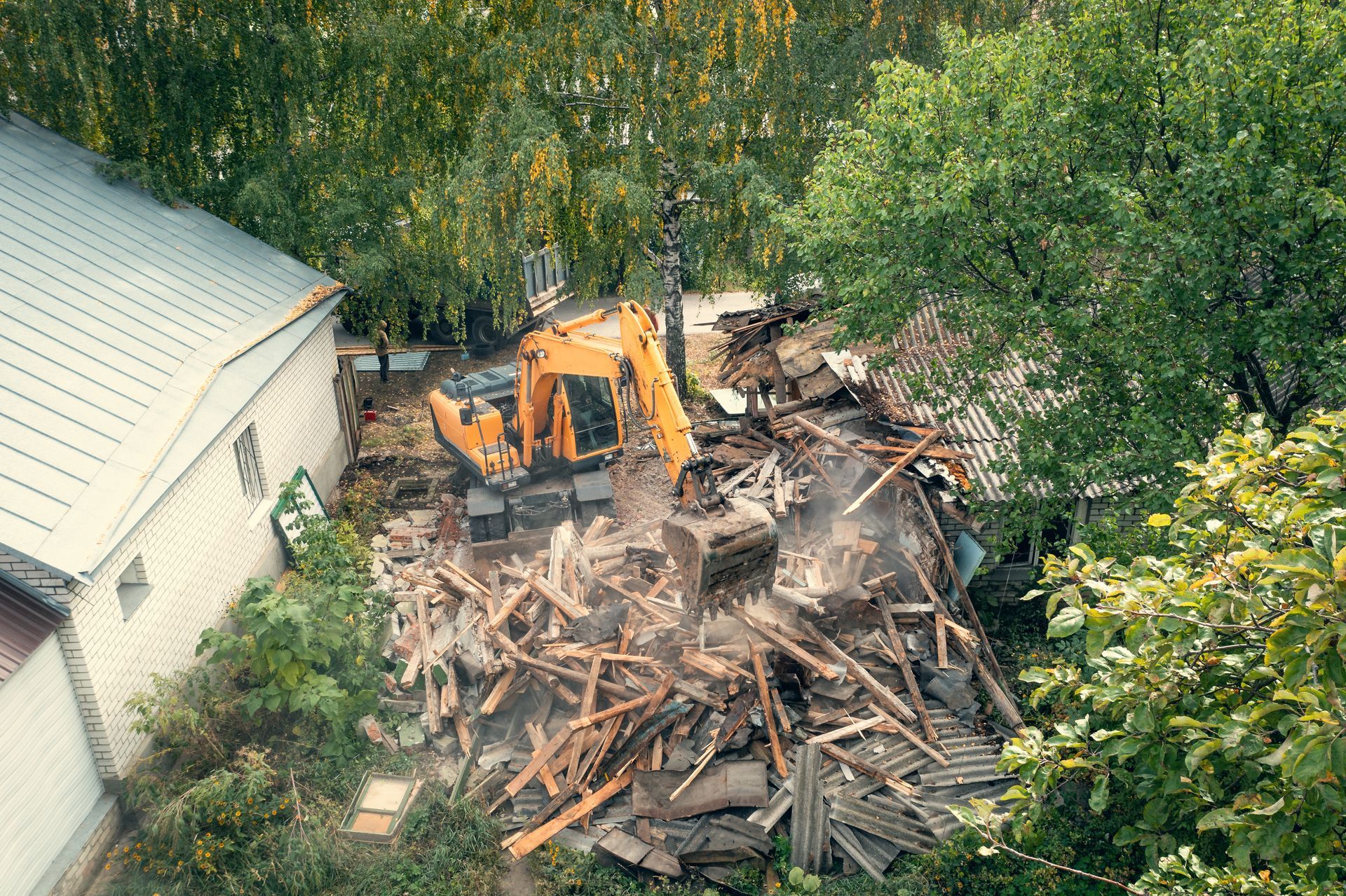 Yellow excavator demolishing a building surrounded by trees. Debris and dust fill the air.