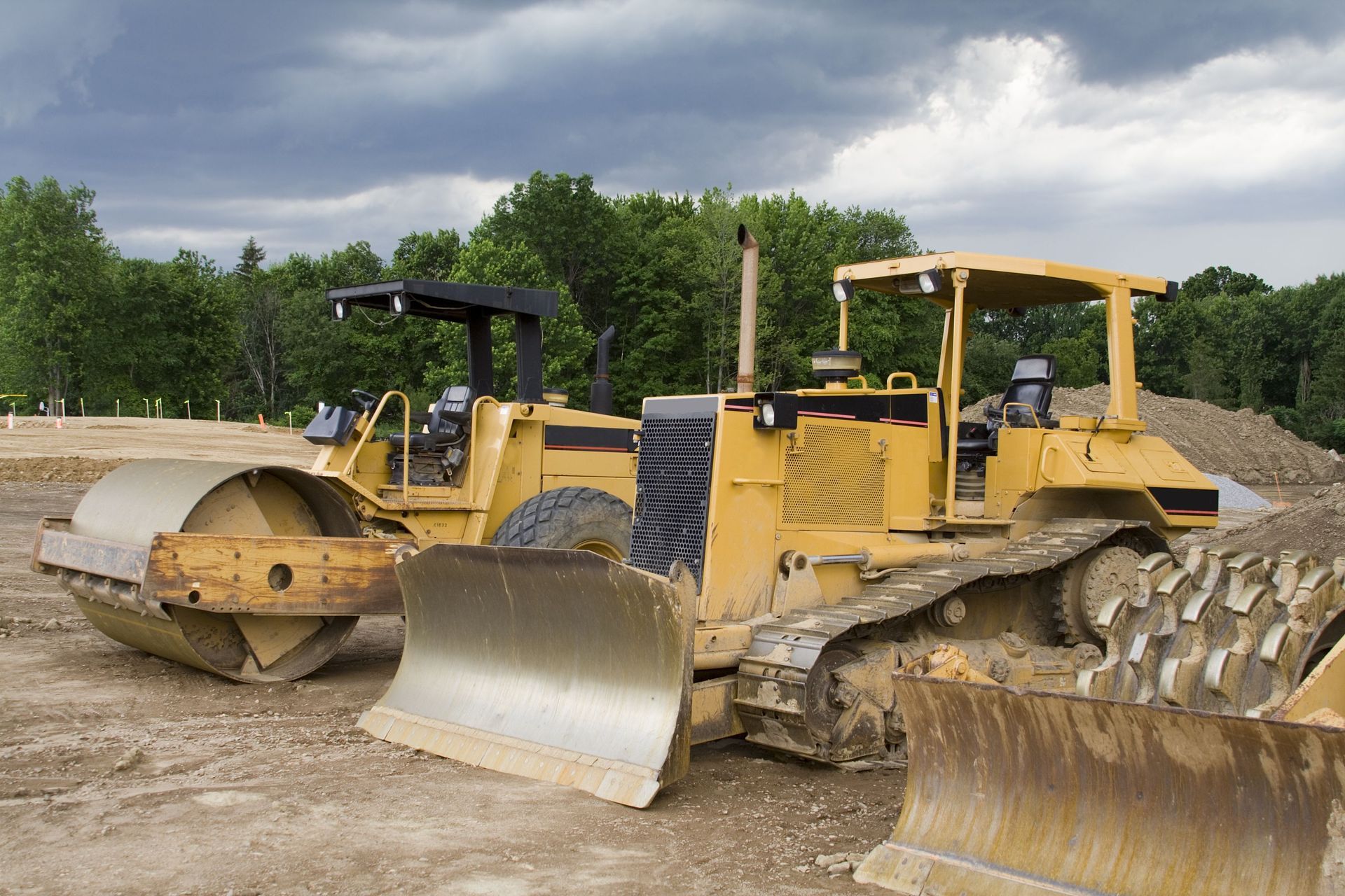 Yellow bulldozer and road roller on construction site. Cloudy sky and trees in the background.