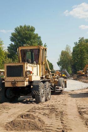 Yellow road grader on a construction site grading gravel, with other machinery in the background.