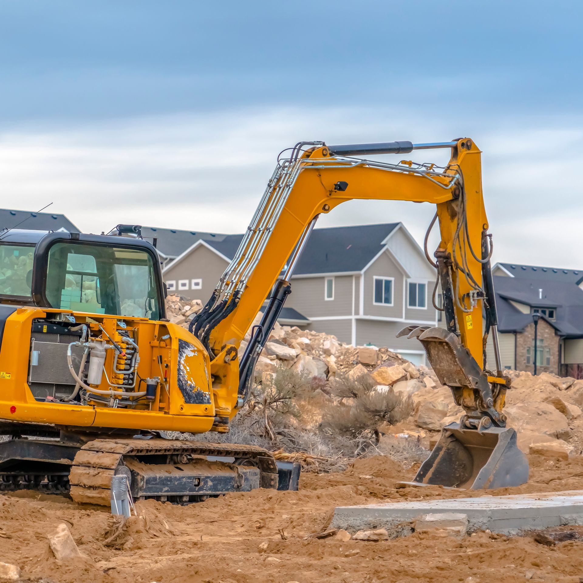 Yellow excavator on a construction site, scooping dirt. Houses in background under a cloudy sky.