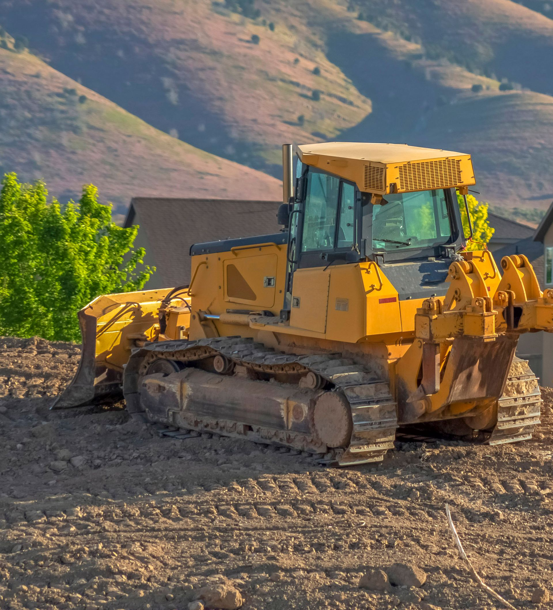 Yellow bulldozer on dirt, with mountains and houses in the background.