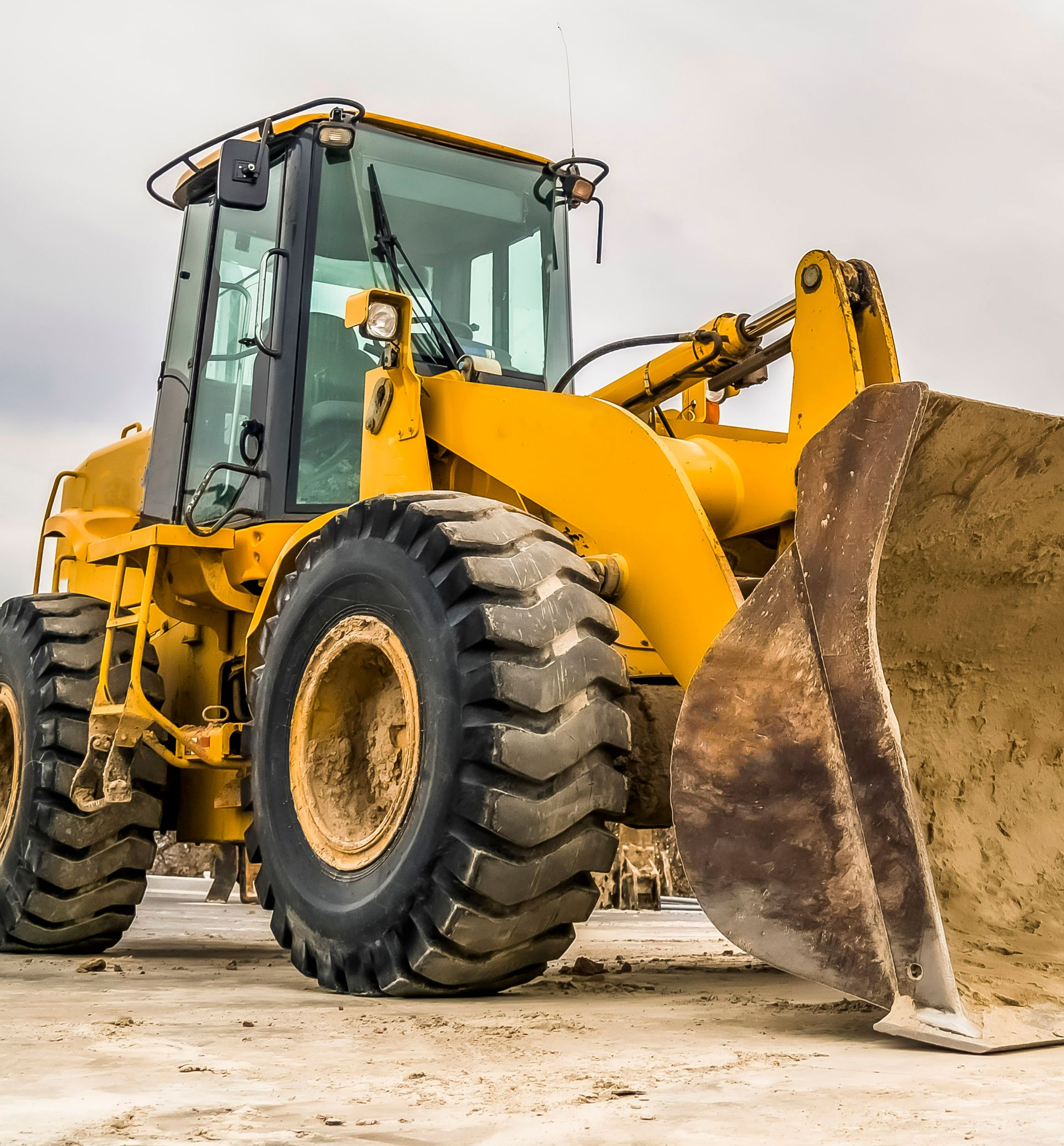 Yellow wheel loader on a construction site, filled scoop, gray overcast sky.