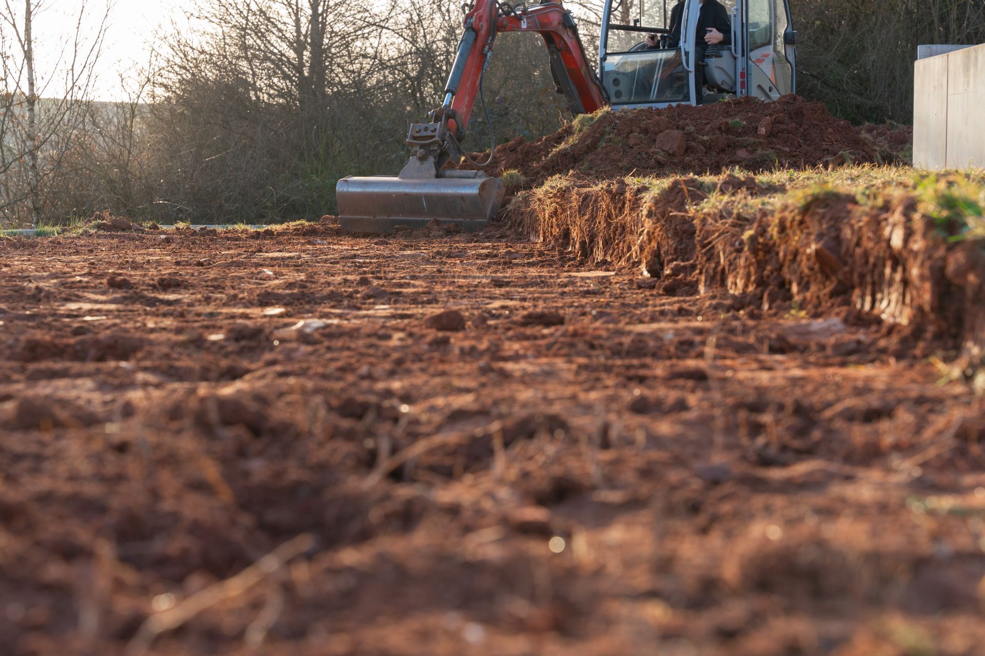 Mini excavator digging a trench in red soil; operator visible in the cab.