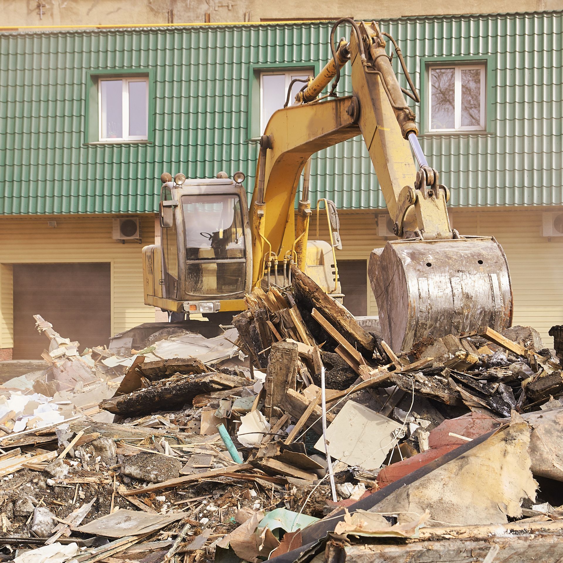 Yellow excavator demolishing a building with green roof and beige walls.