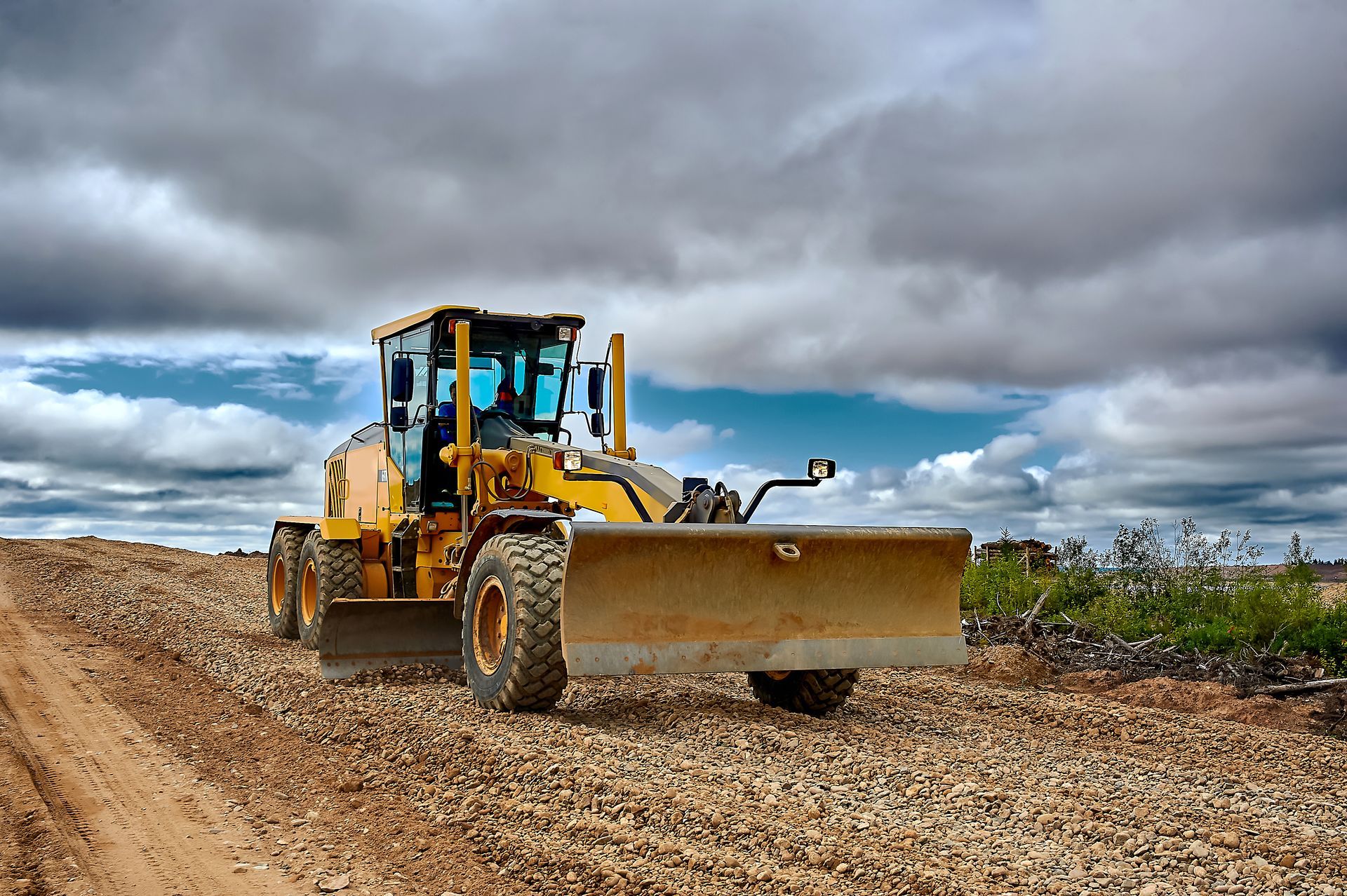 Yellow motor grader on a rocky road, under a cloudy sky.
