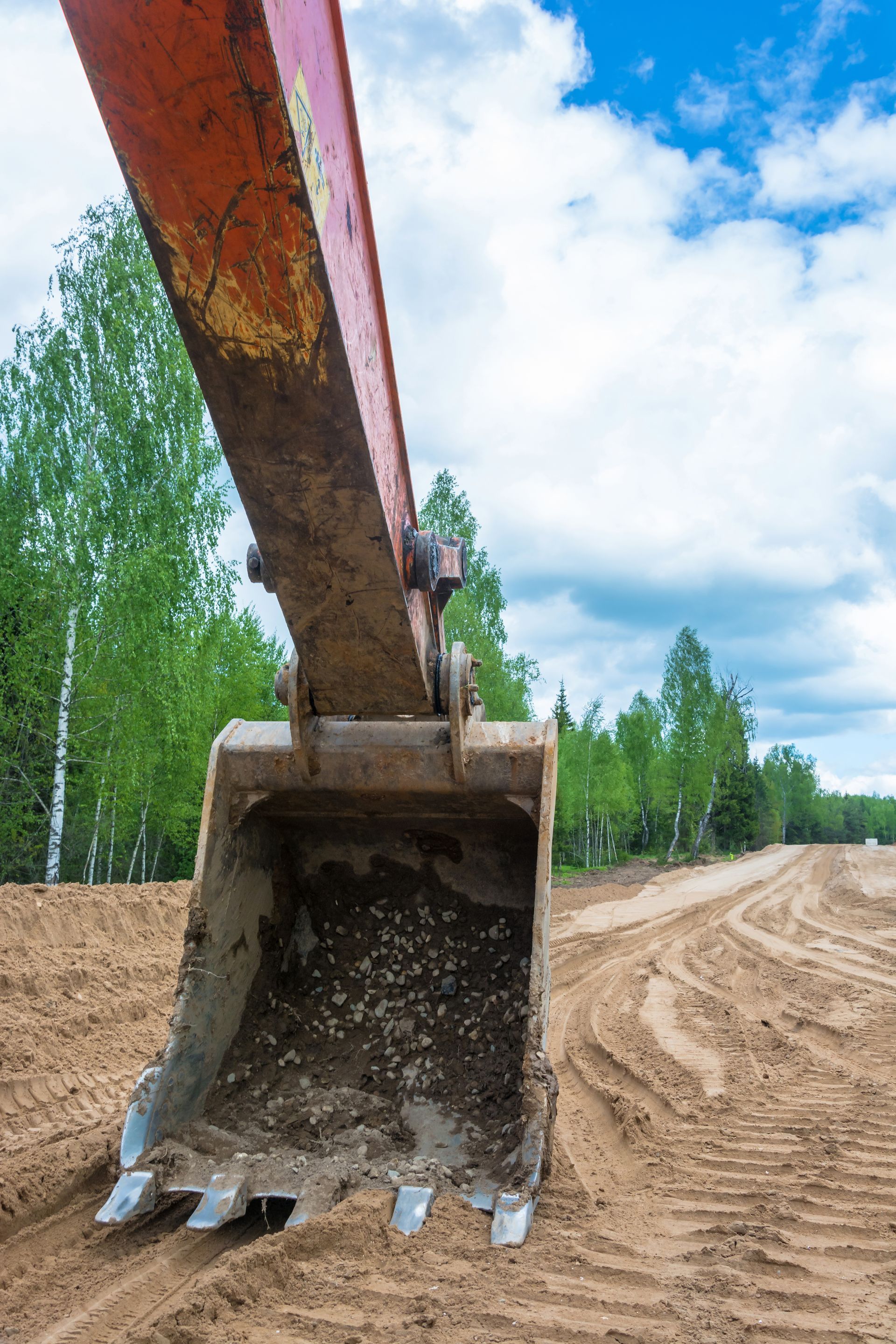 Excavator bucket scooping sand on a construction site, blue sky, green trees in background.