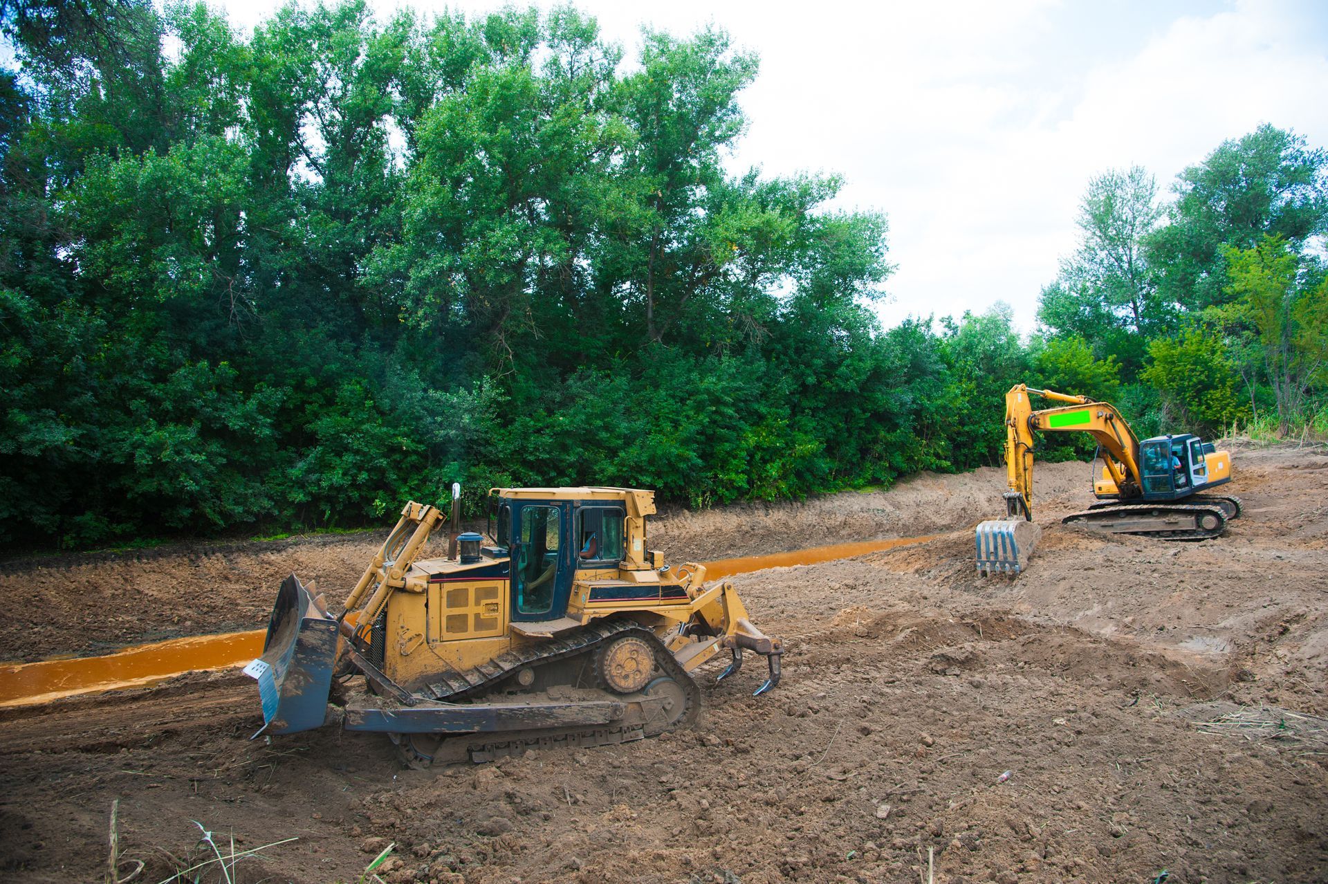 Two construction vehicles, a bulldozer and an excavator, working on muddy terrain near a line of trees.