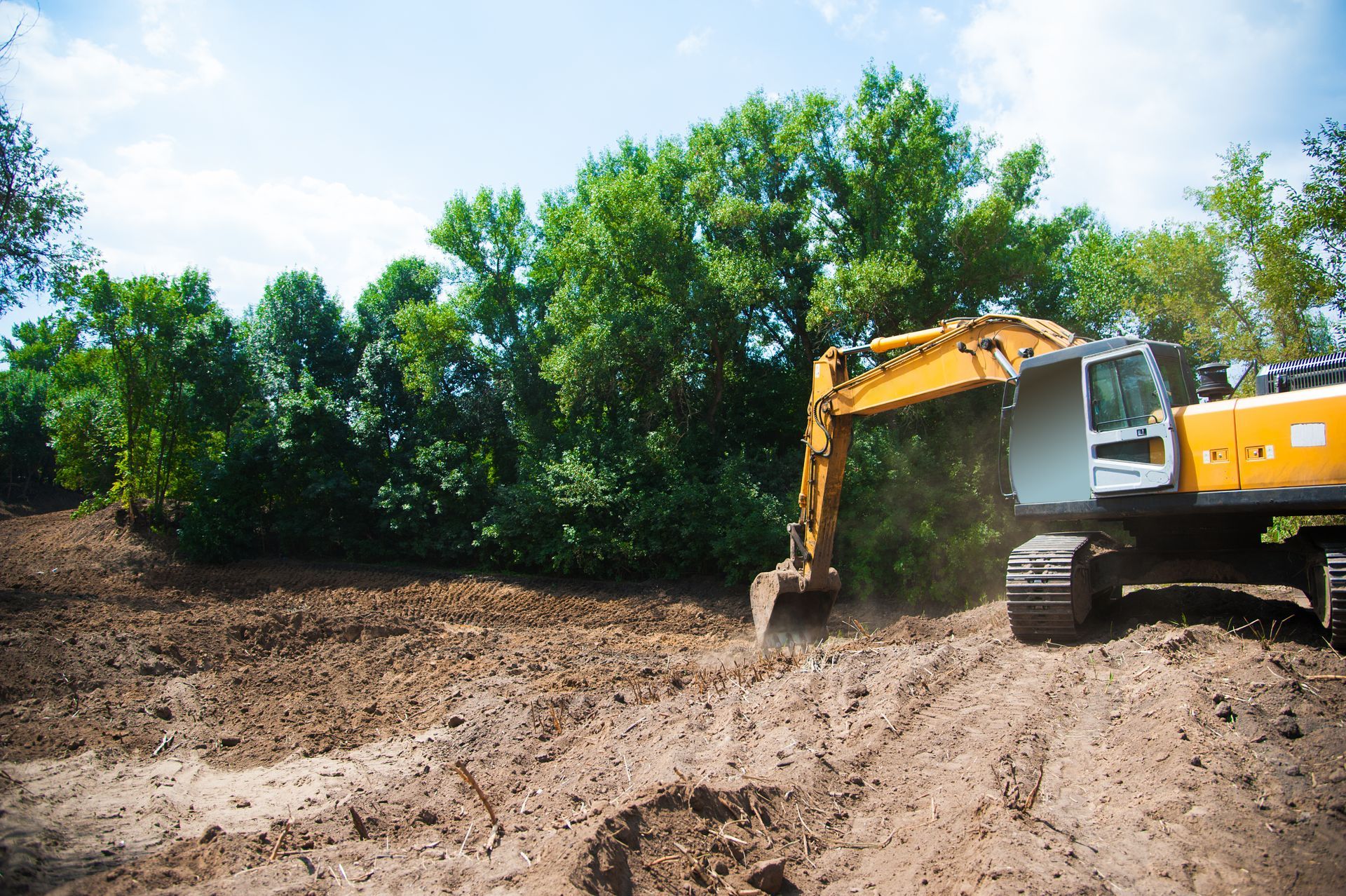 Yellow excavator digging in muddy field, trees in the background under blue sky.