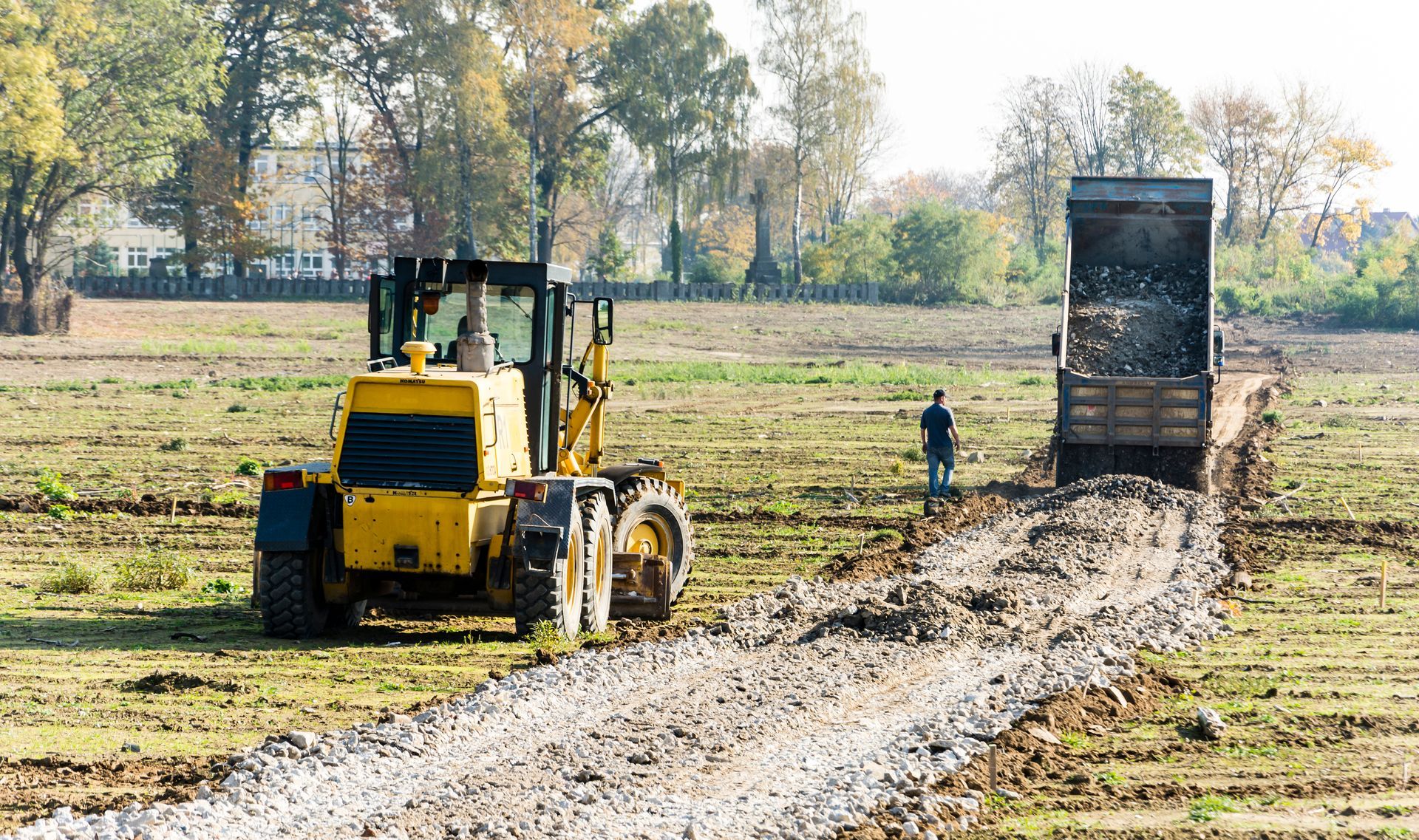 Yellow construction vehicle and dump truck laying gravel path in field; person nearby.