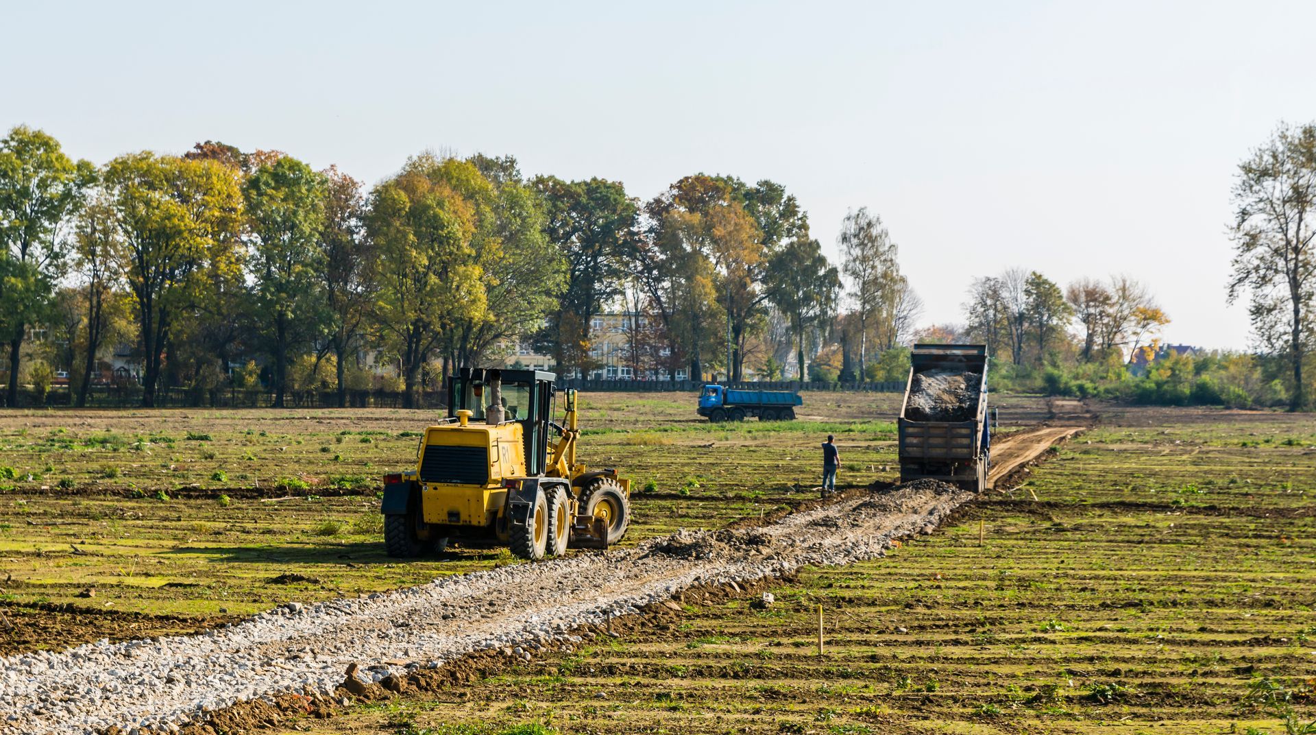 Yellow tractor and dump truck on construction site, trees in background.
