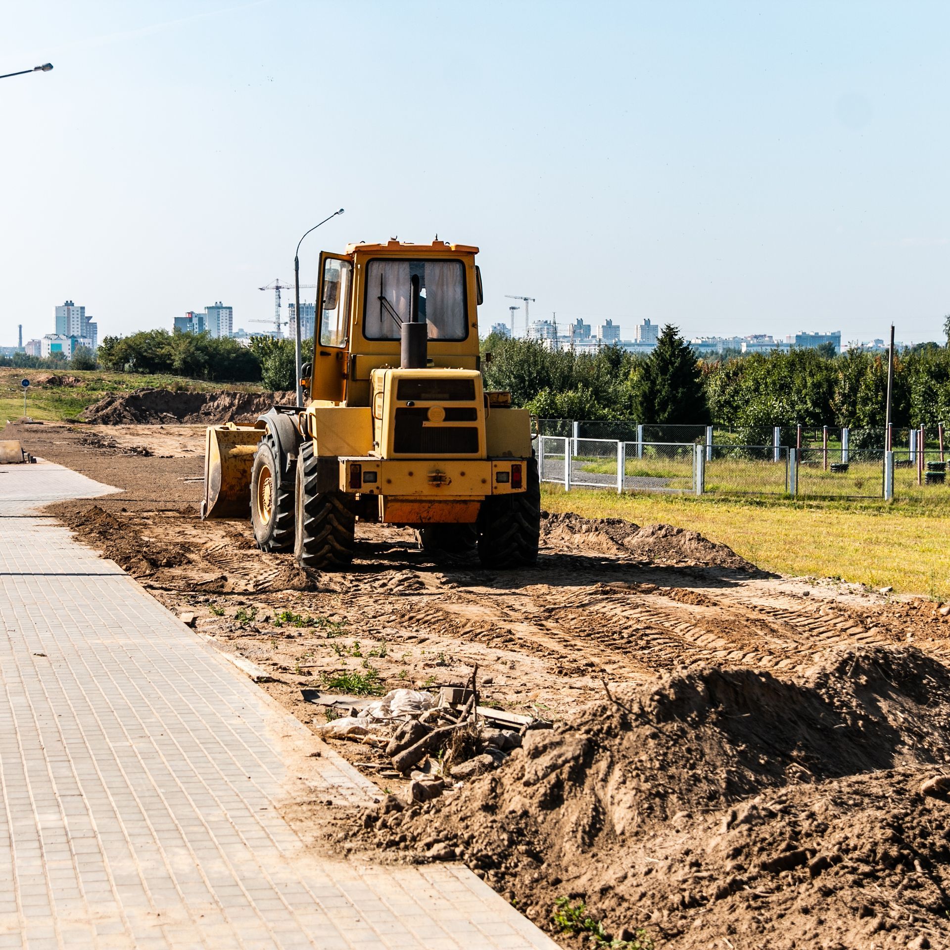 Yellow construction tractor on a dirt path next to a brick-paved sidewalk with a city skyline in the background.