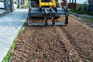 A construction machine with a claw-like attachment clearing debris from a driveway, next to a paved walkway.