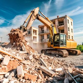 Yellow excavator demolishing a building with rubble in the foreground under a blue sky.