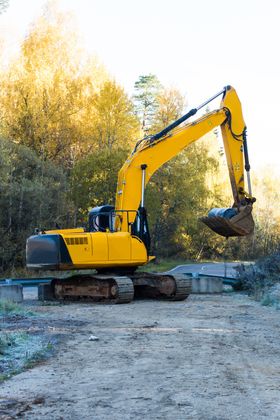 Yellow excavator on a dirt road, arm raised. Forest background with golden foliage.