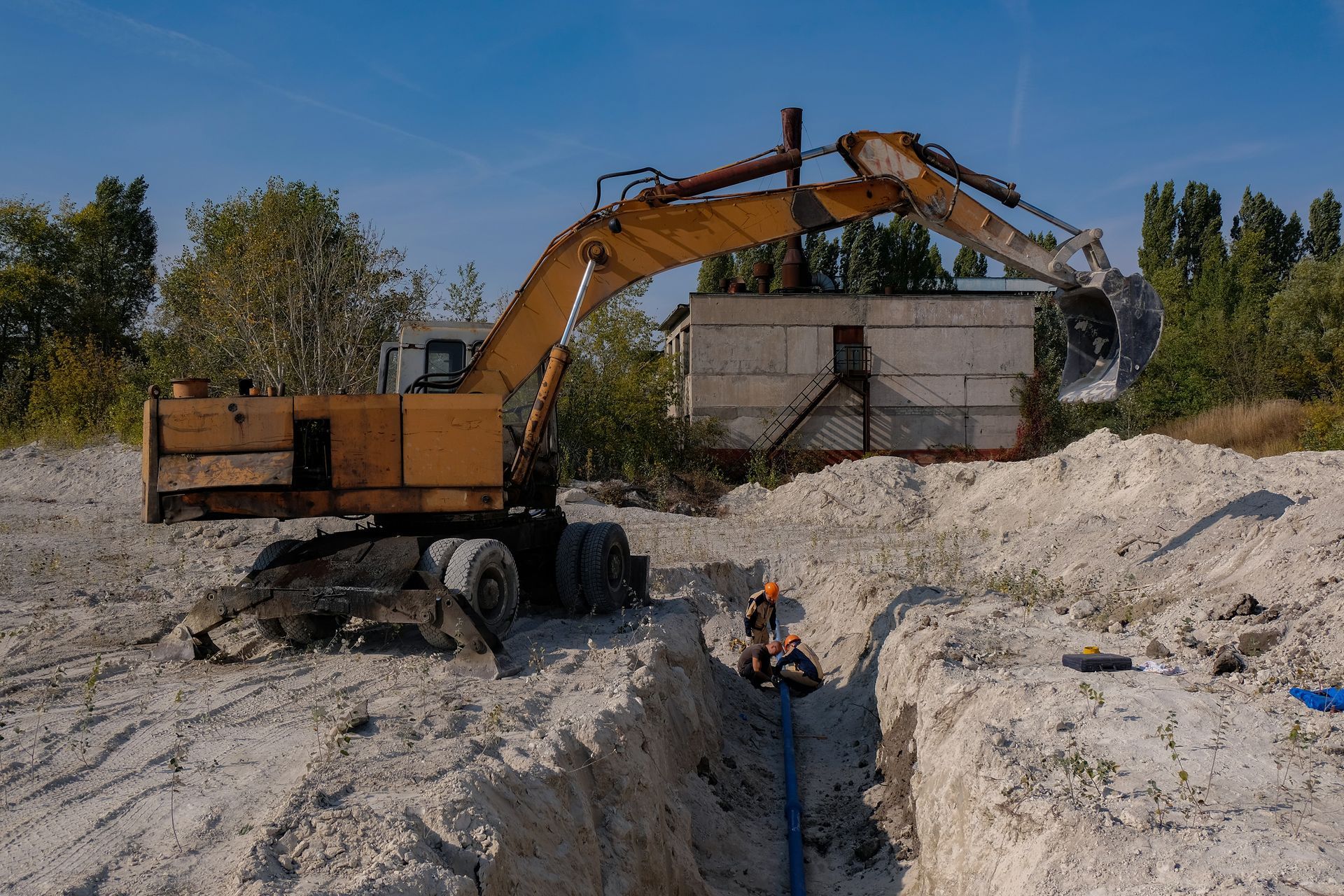 Yellow excavator over a trench, worker in the trench, white dirt, blue pipe, small building, trees, and a clear blue sky.