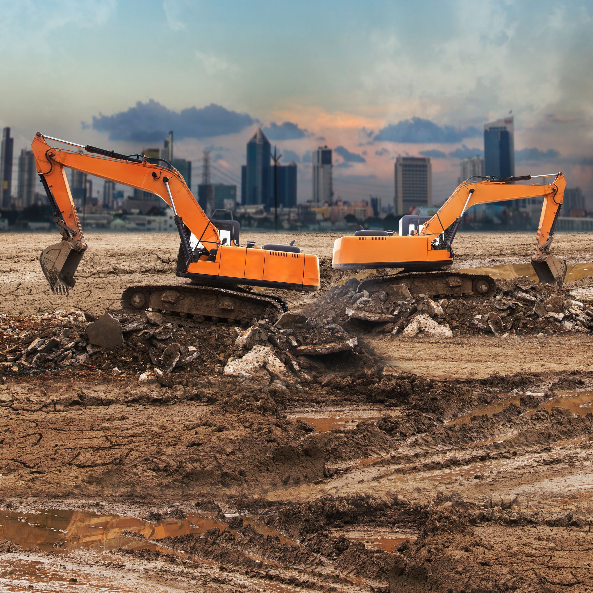 Two orange excavators on a construction site with a city skyline backdrop.