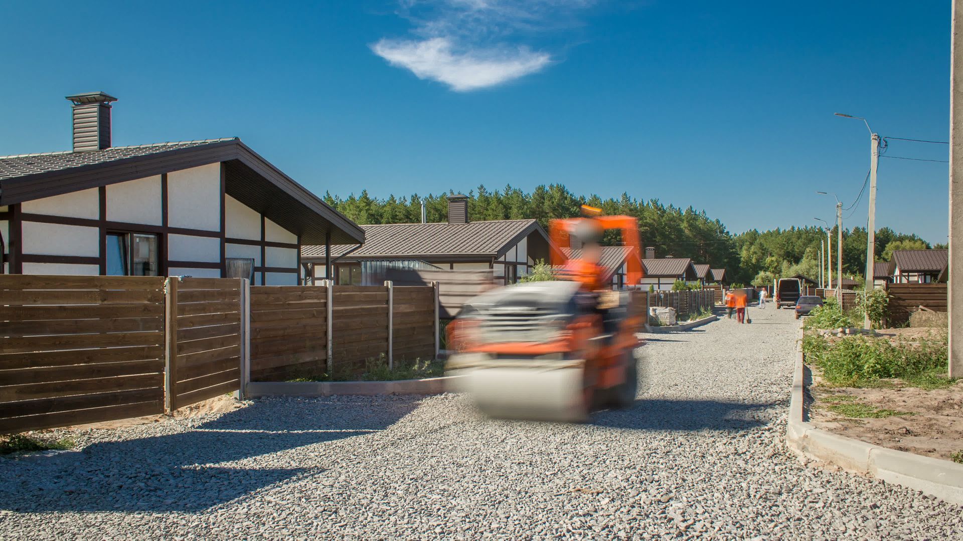Orange road roller compacting gravel road in front of houses on a sunny day.