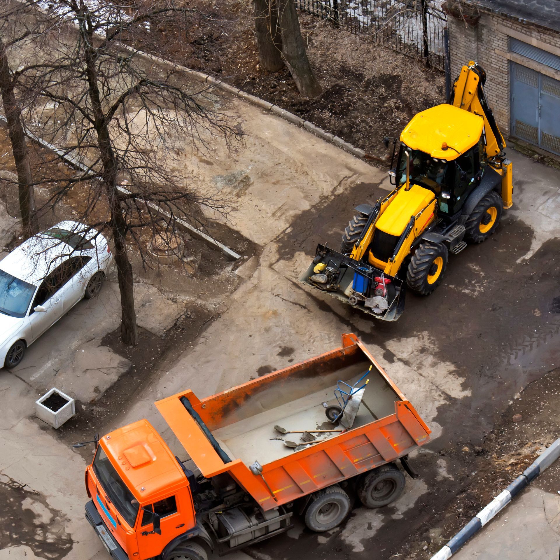 Yellow backhoe loader next to an orange dump truck on a construction site. A white car and trees are also present.