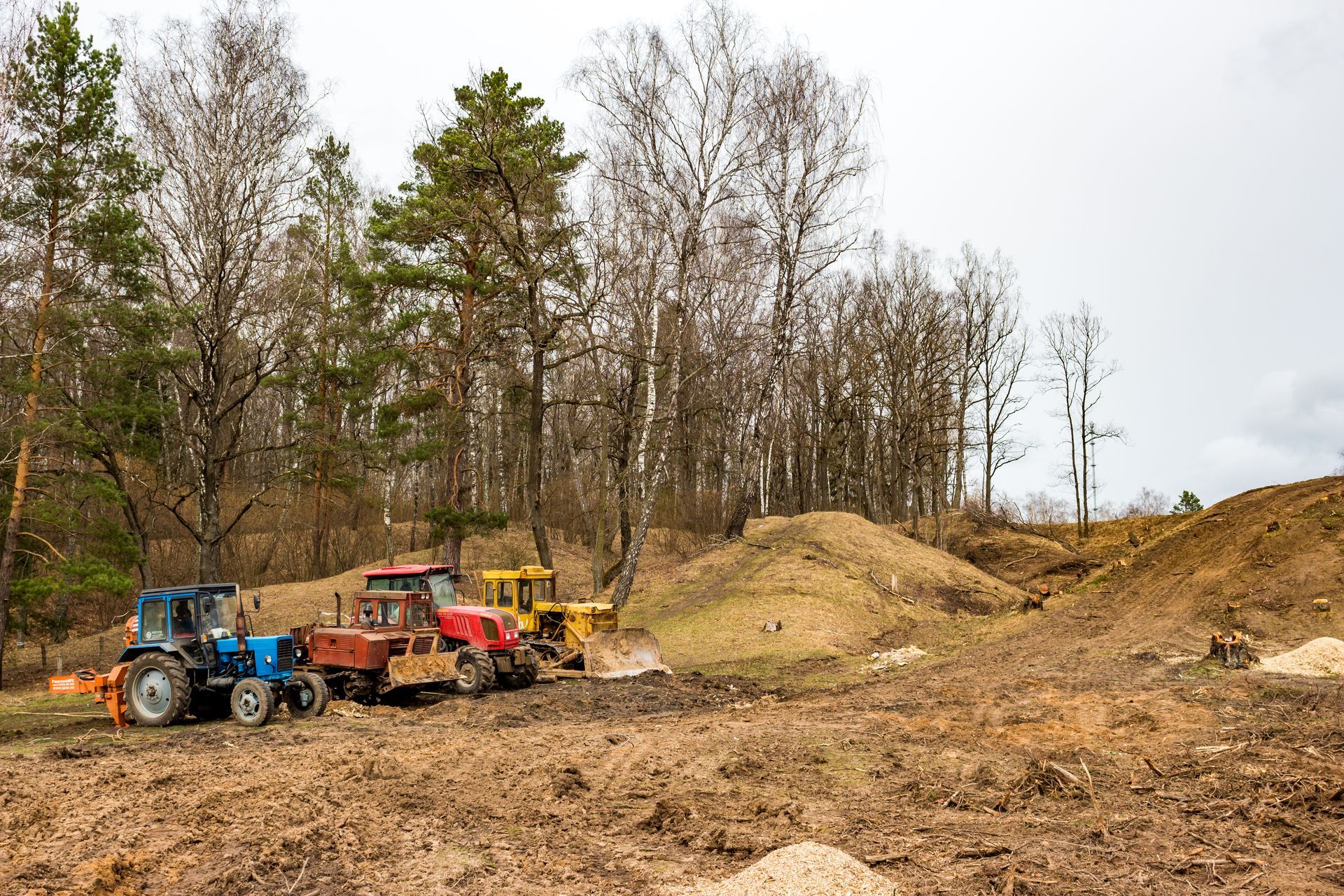Three tractors working on a dirt field near a forest; brown, blue, red, and yellow vehicles.
