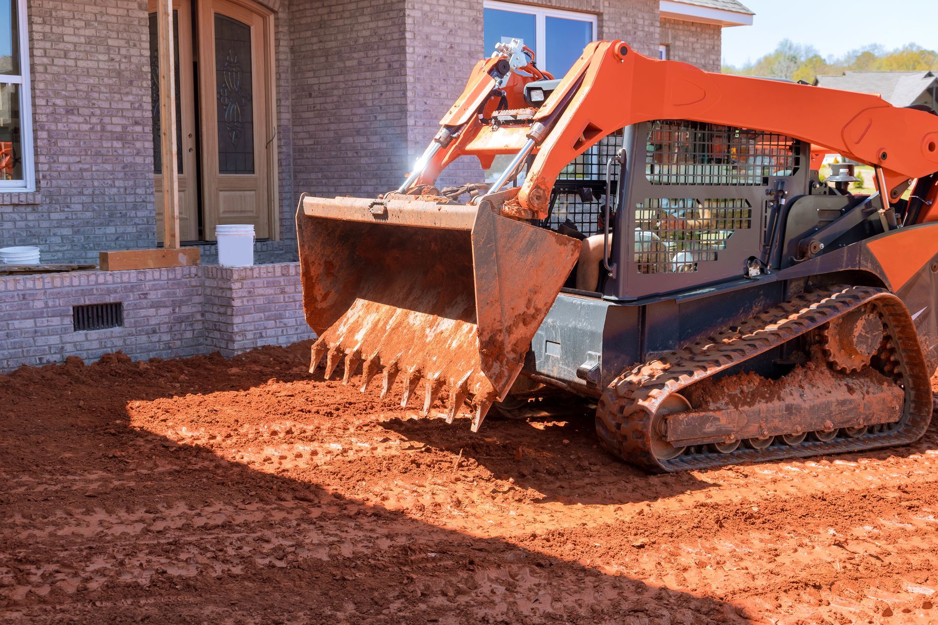 Orange skid steer moving reddish-brown dirt near a brick house under construction.