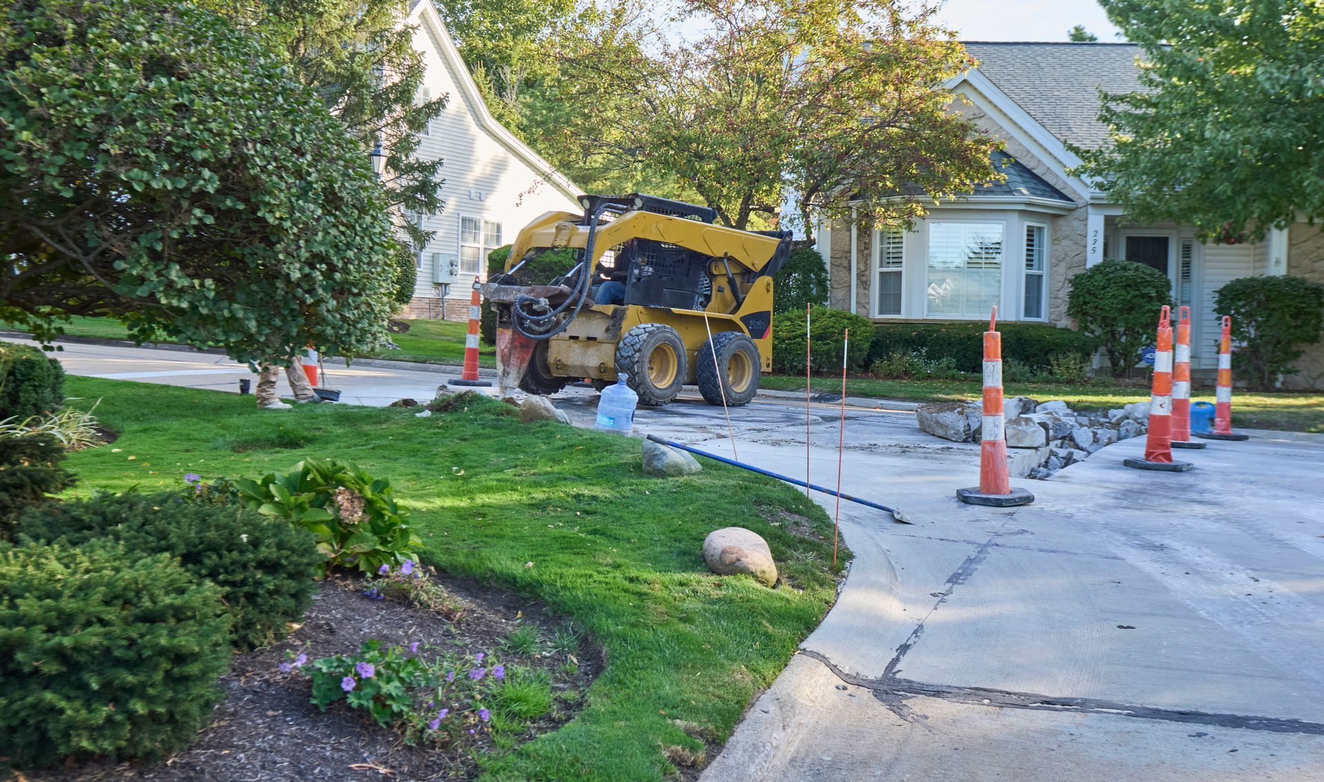 Yellow construction vehicle working on a driveway in a residential area, with orange traffic cones.
