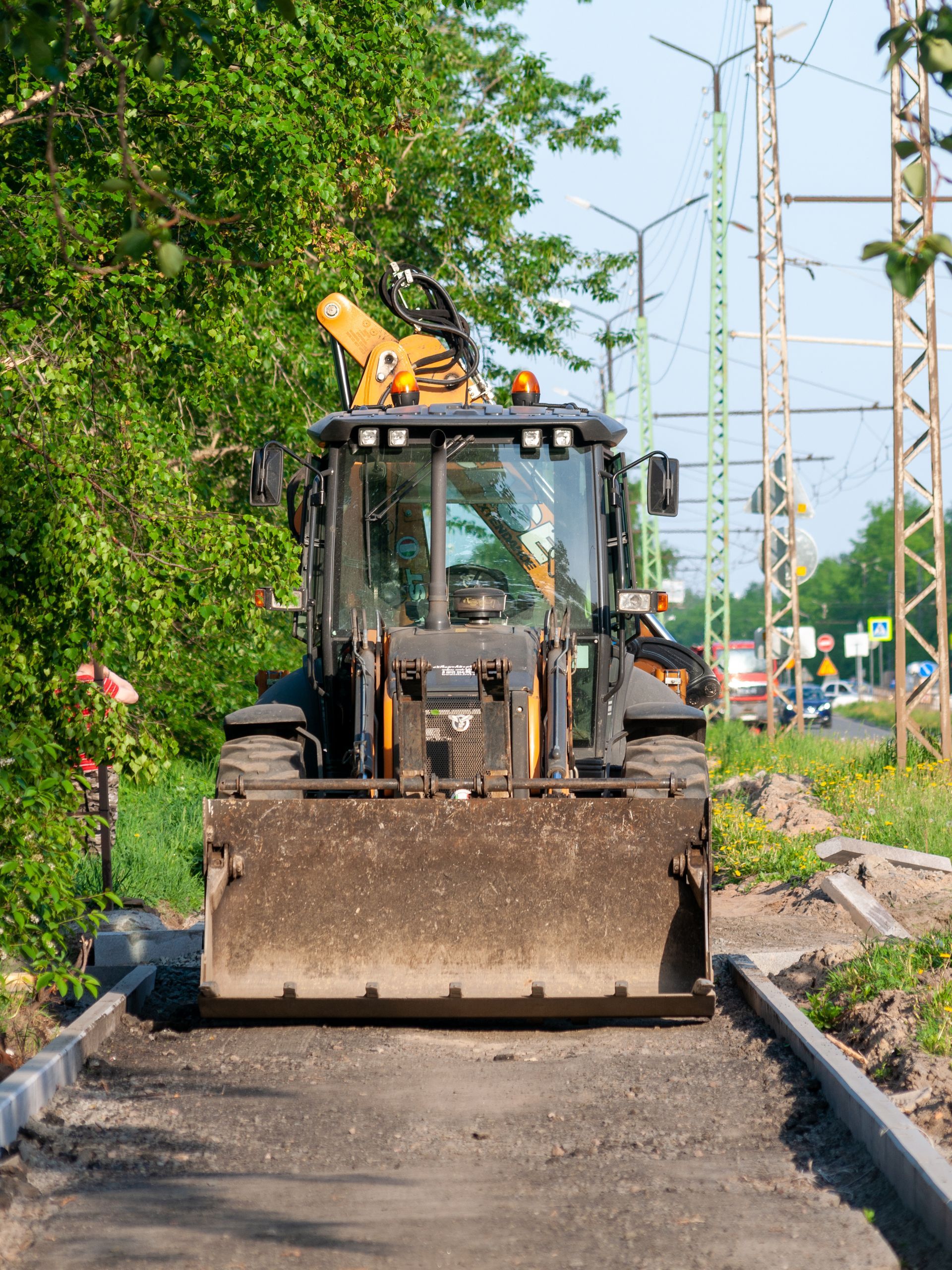 Backhoe on asphalt path, preparing for road work, near a green tree and road.