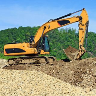Yellow excavator digging dirt on a construction site with green trees in the background.