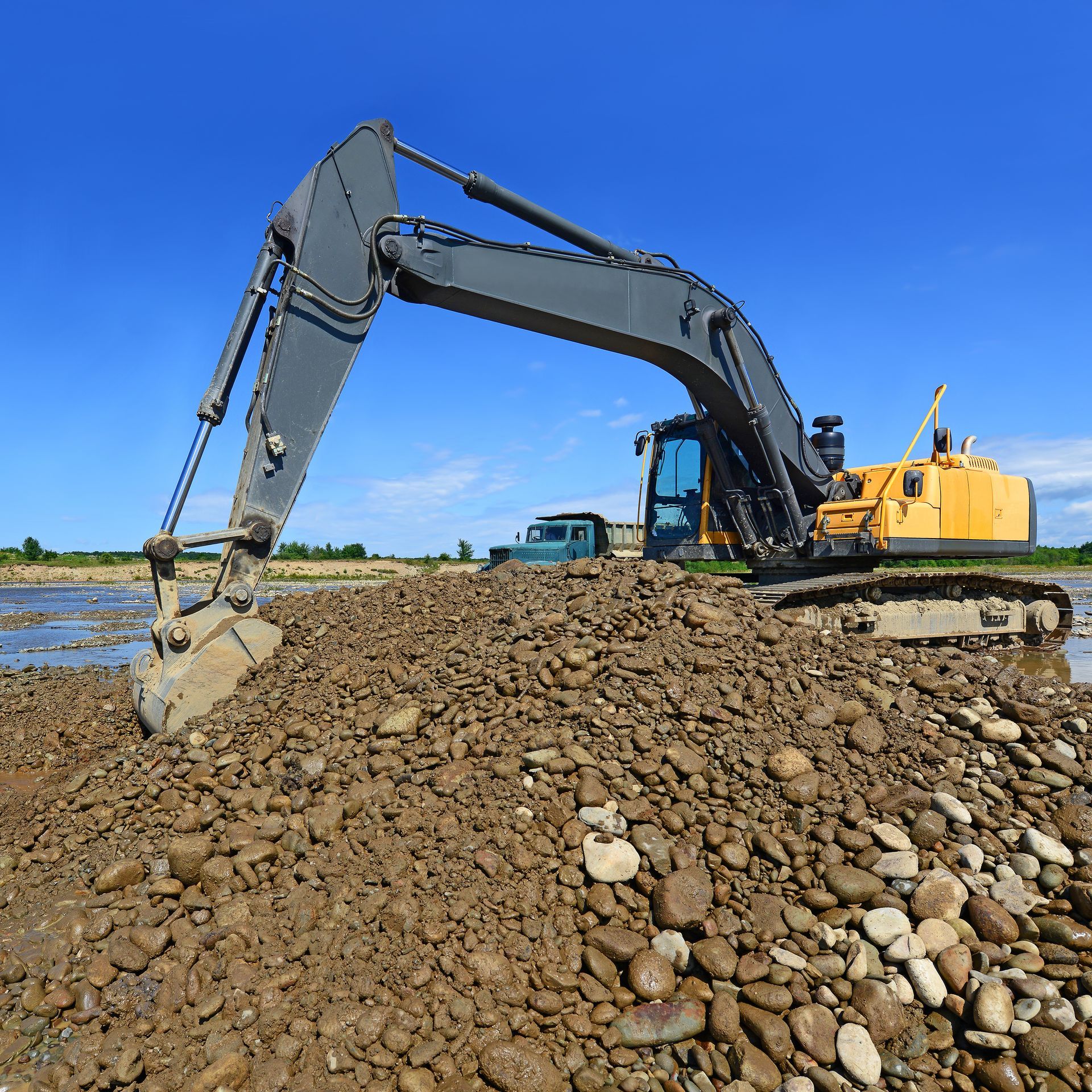 Excavator on a pile of dirt and rocks under a blue sky, near a body of water and a parked truck.